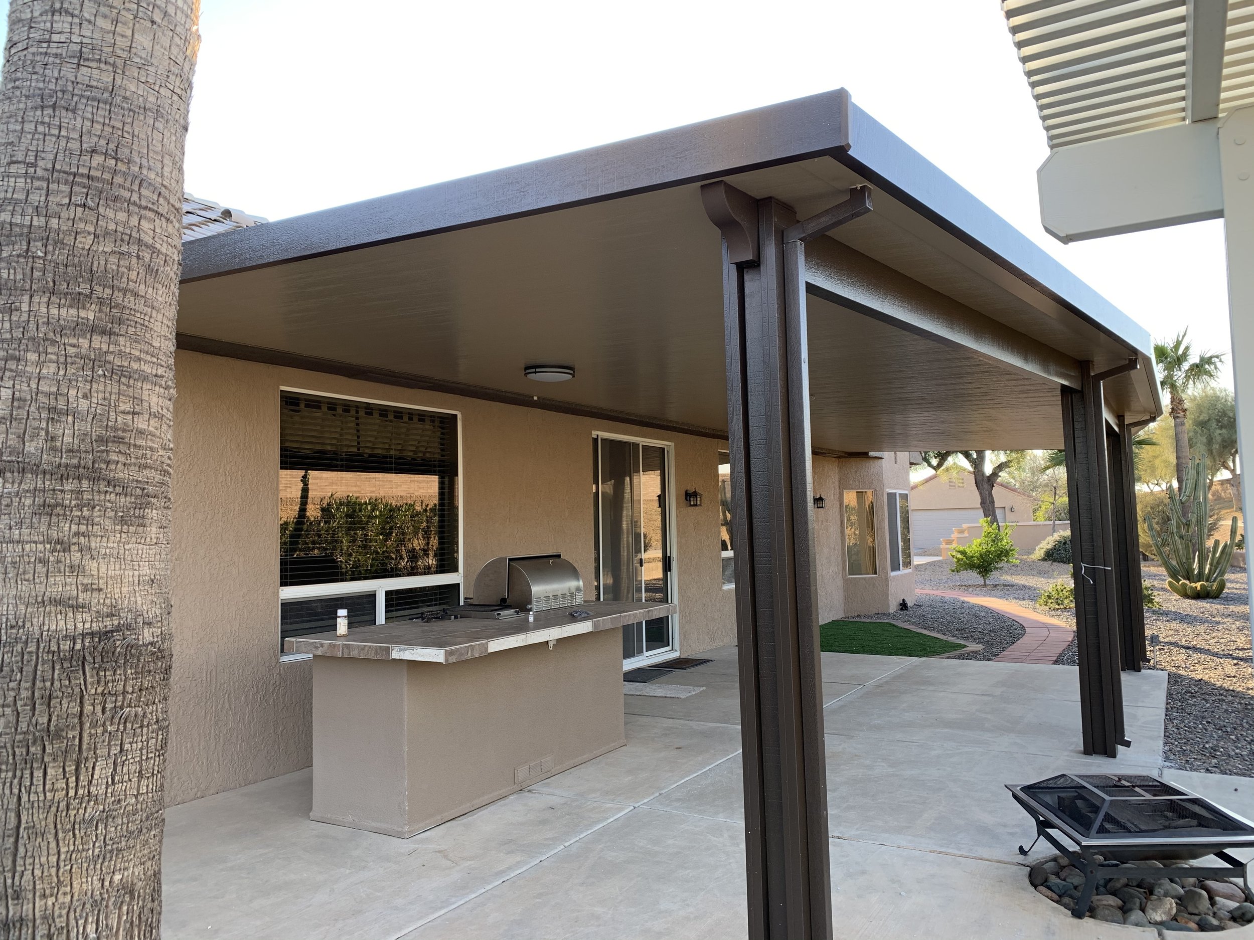 View of a backyard patio with a covered area, outdoor grill on a concrete counter, and desert landscaping with cacti and small trees.