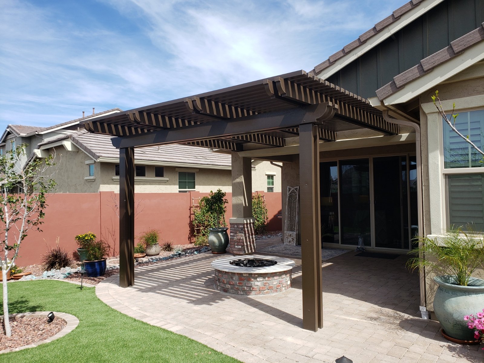 Backyard patio area with a wooden pergola, brick fire pit, potted plants, and a sliding glass door leading into the house. The yard has a small patch of artificial grass and landscaped garden beds.