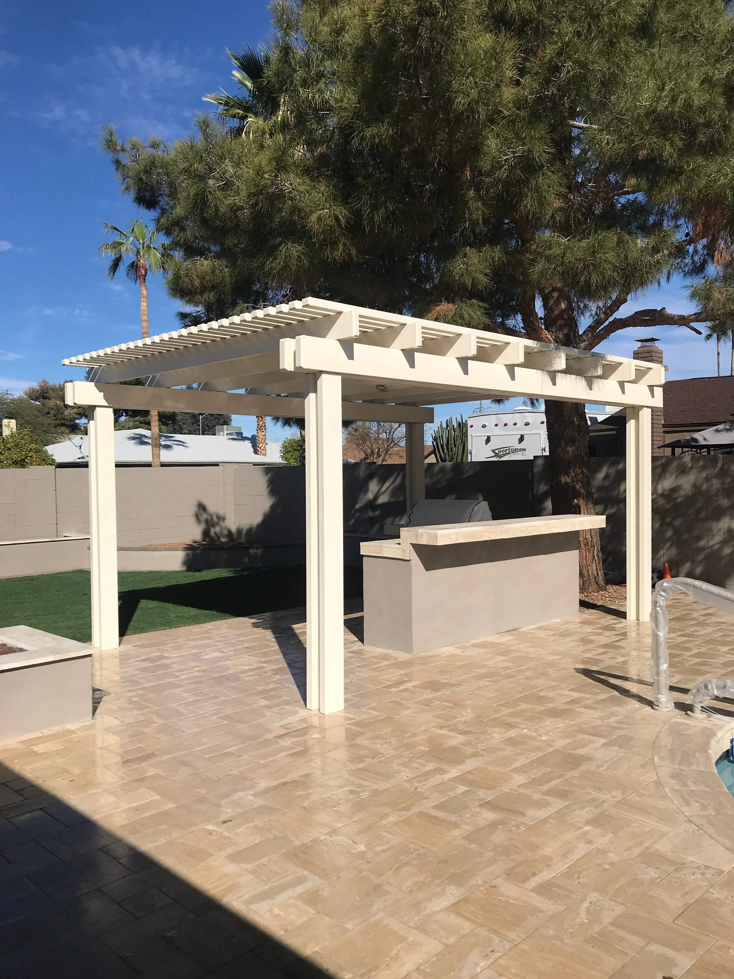Backyard patio with a white pergola, a built-in outdoor bar, and a swimming pool, with trees and a gray fence in the background.