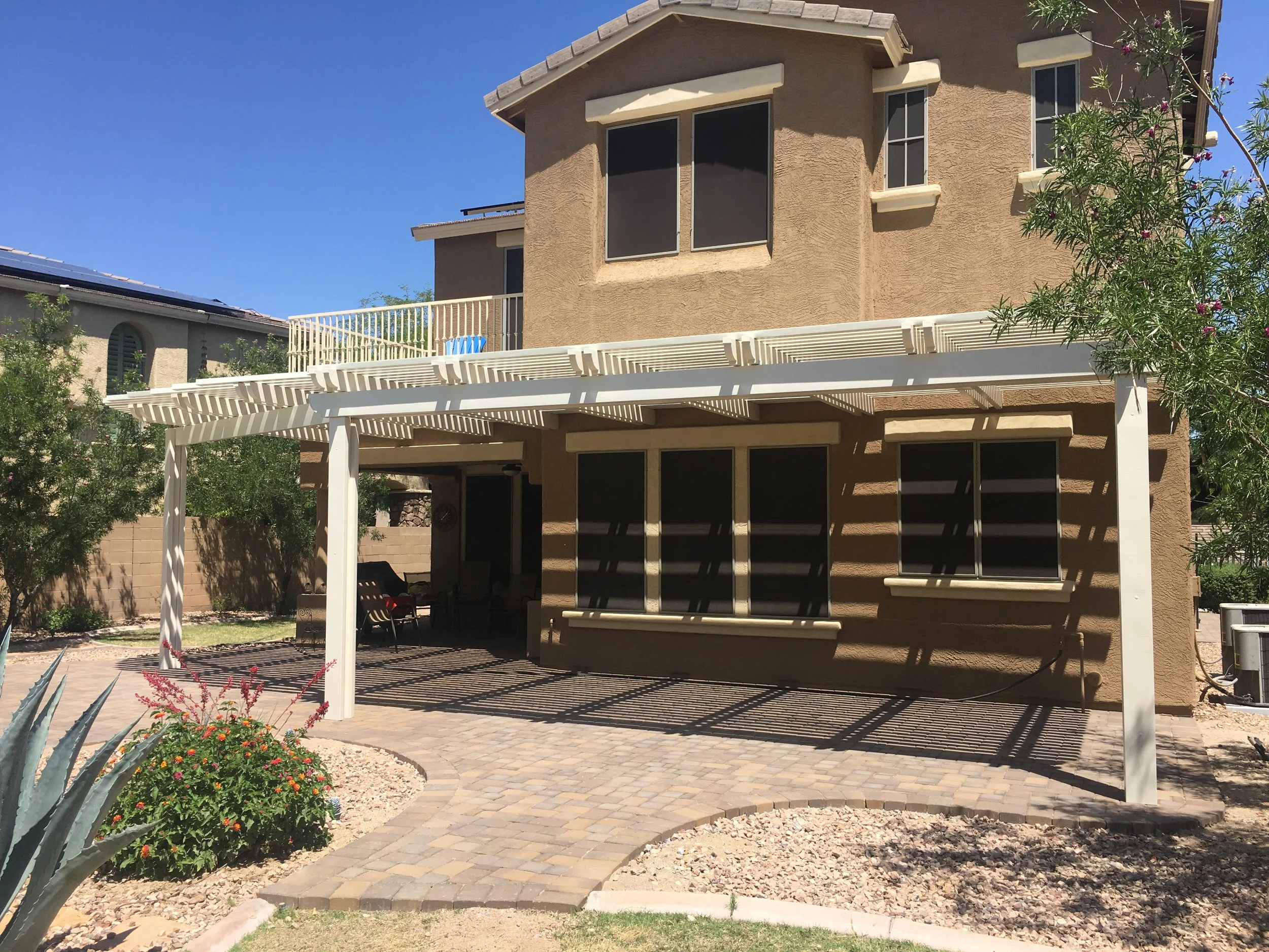 Backyard with a beige two-story house, white pergola, brick patio, and landscaped yard with plants and flowers.