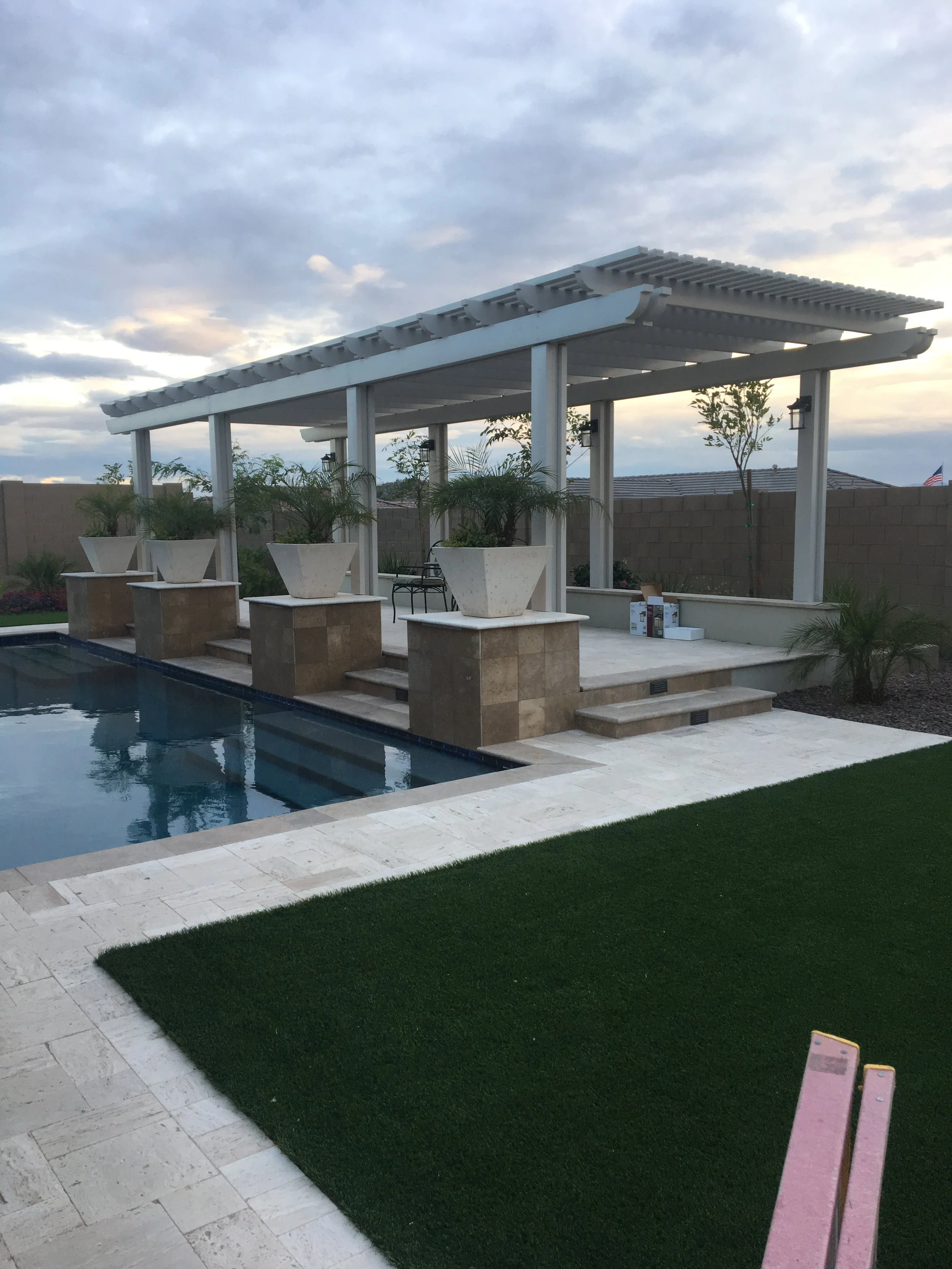 Backyard with swimming pool, white pergola, large potted plants, and a grassy area in the evening with cloudy sky.