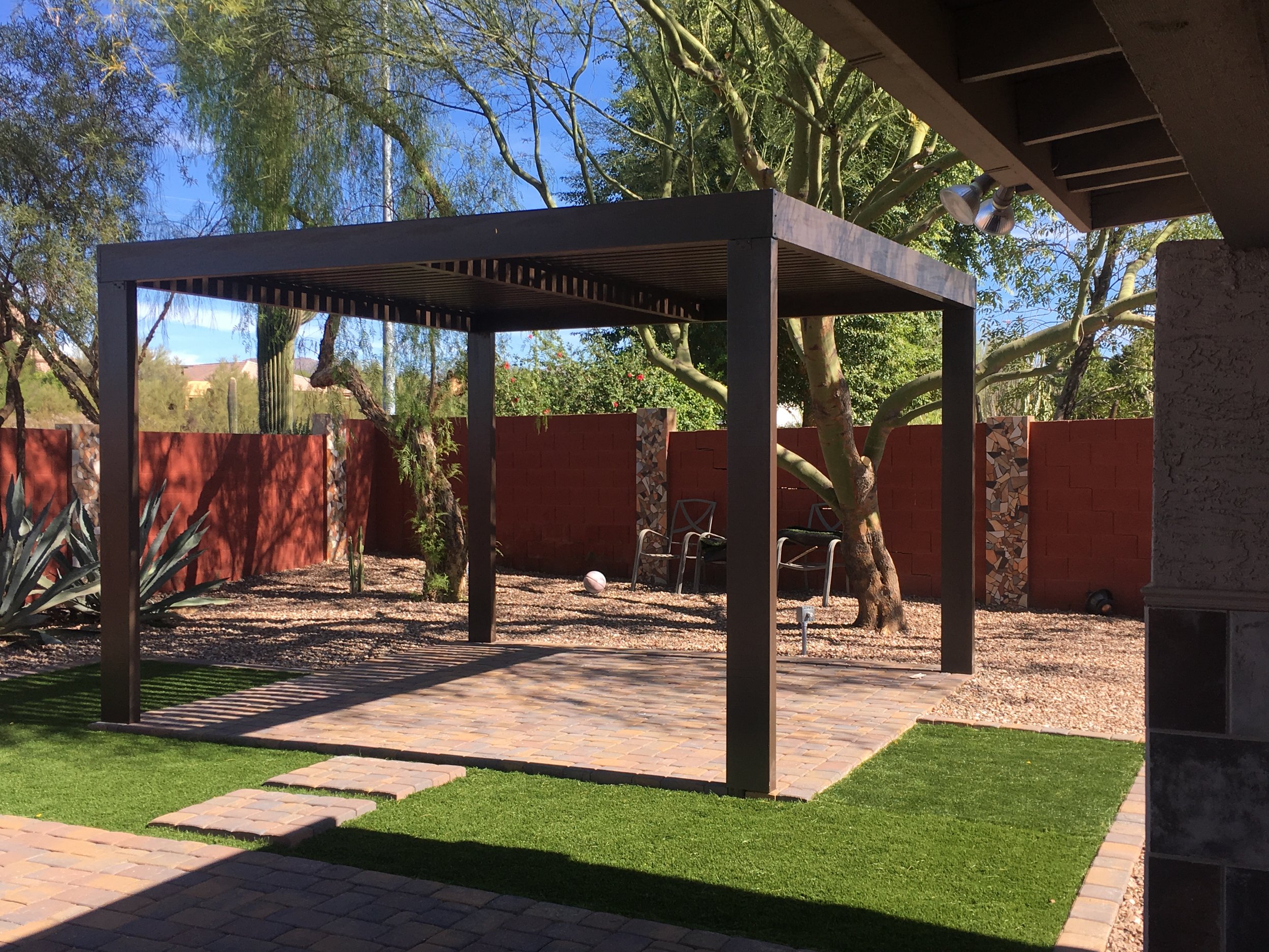Backyard patio with a black pergola, brick pathway, green grass, a red brick wall, desert plants, and trees under a clear blue sky.