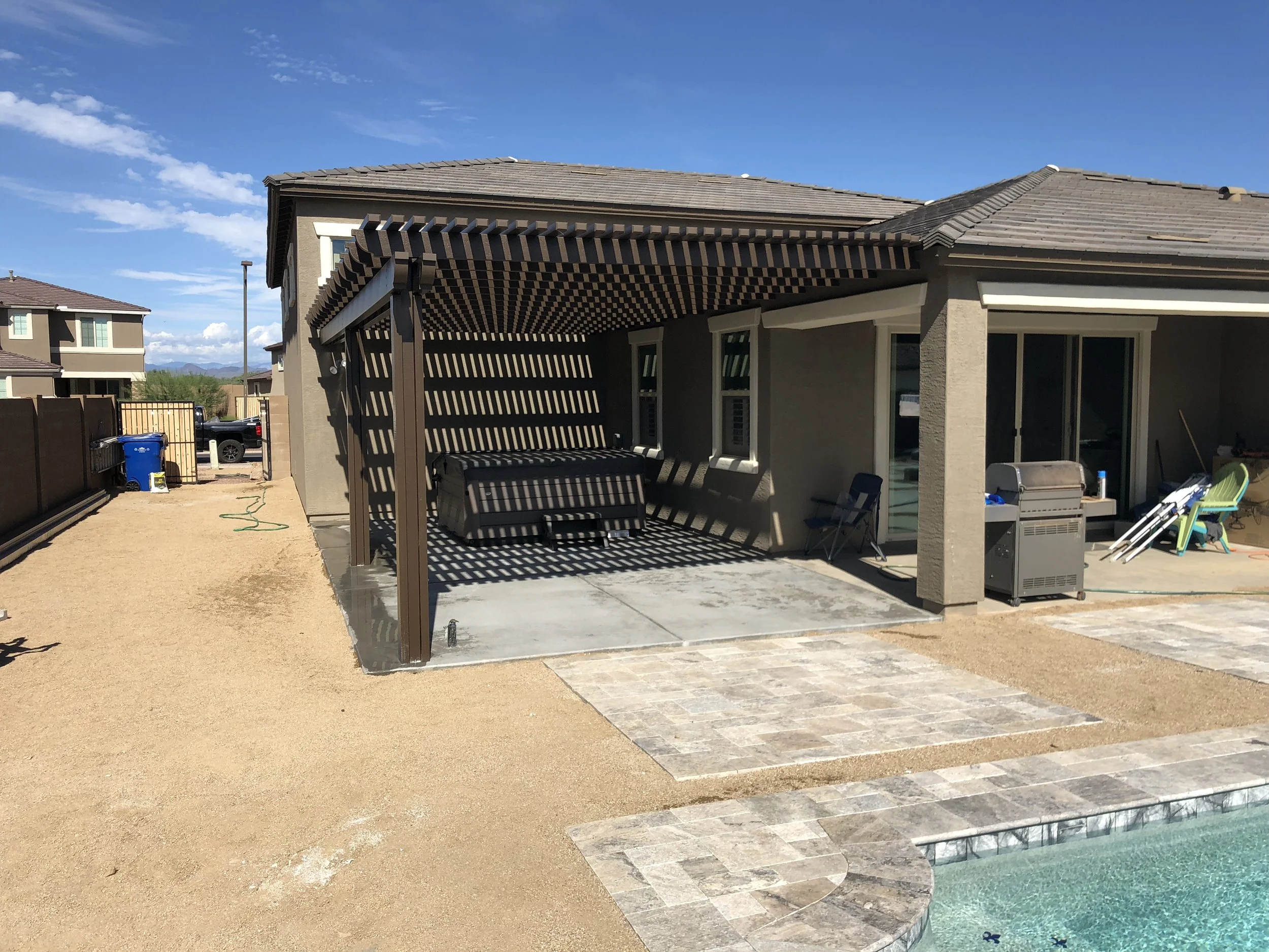 Backyard patio with outdoor seating, grill, and shade structure near a swimming pool, fenced yard, neighboring houses, and clear blue sky.