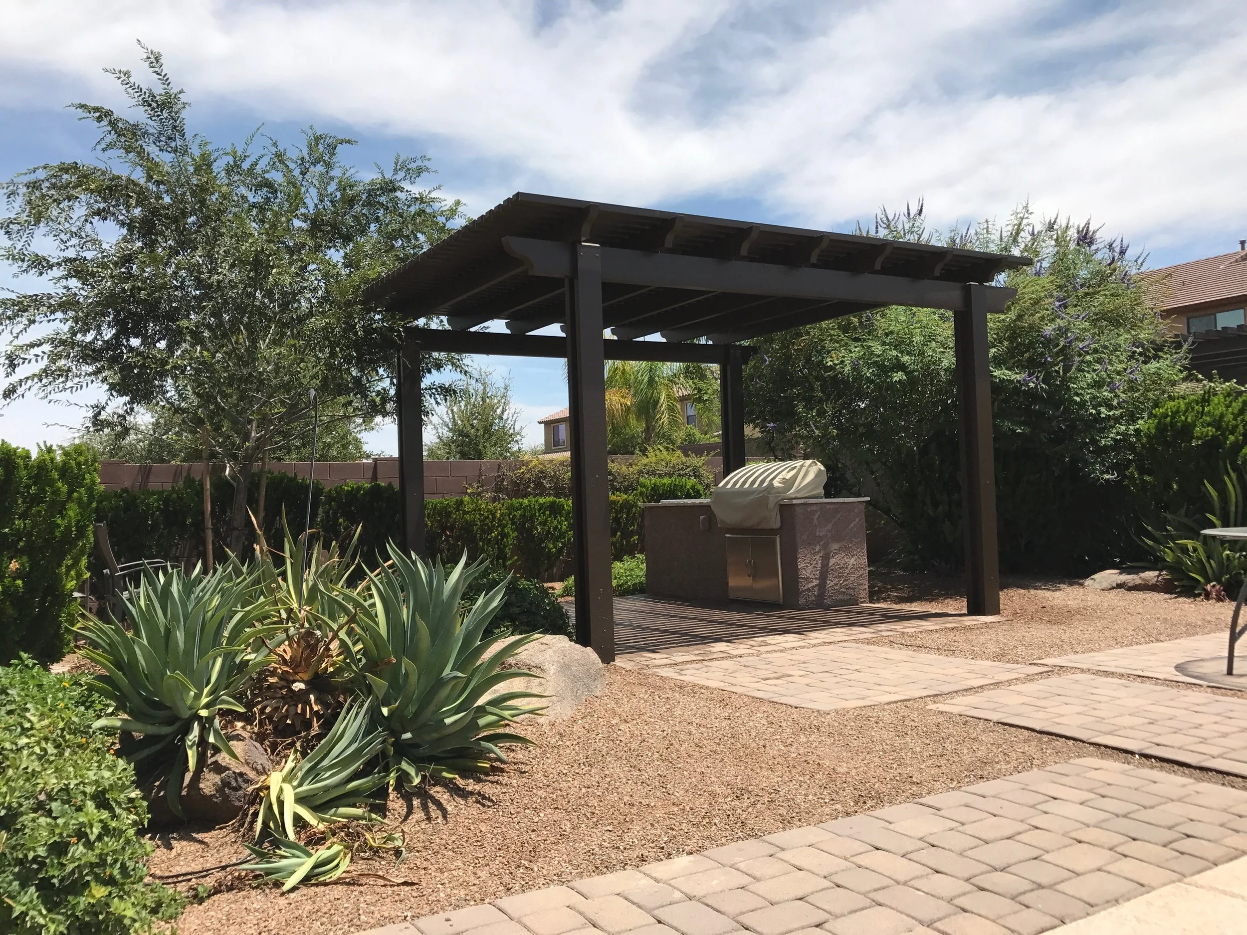 A small outdoor patio area with a covered barbecue grill station, surrounded by desert plants and shrubs, and a paved walkway, under a partly cloudy sky.