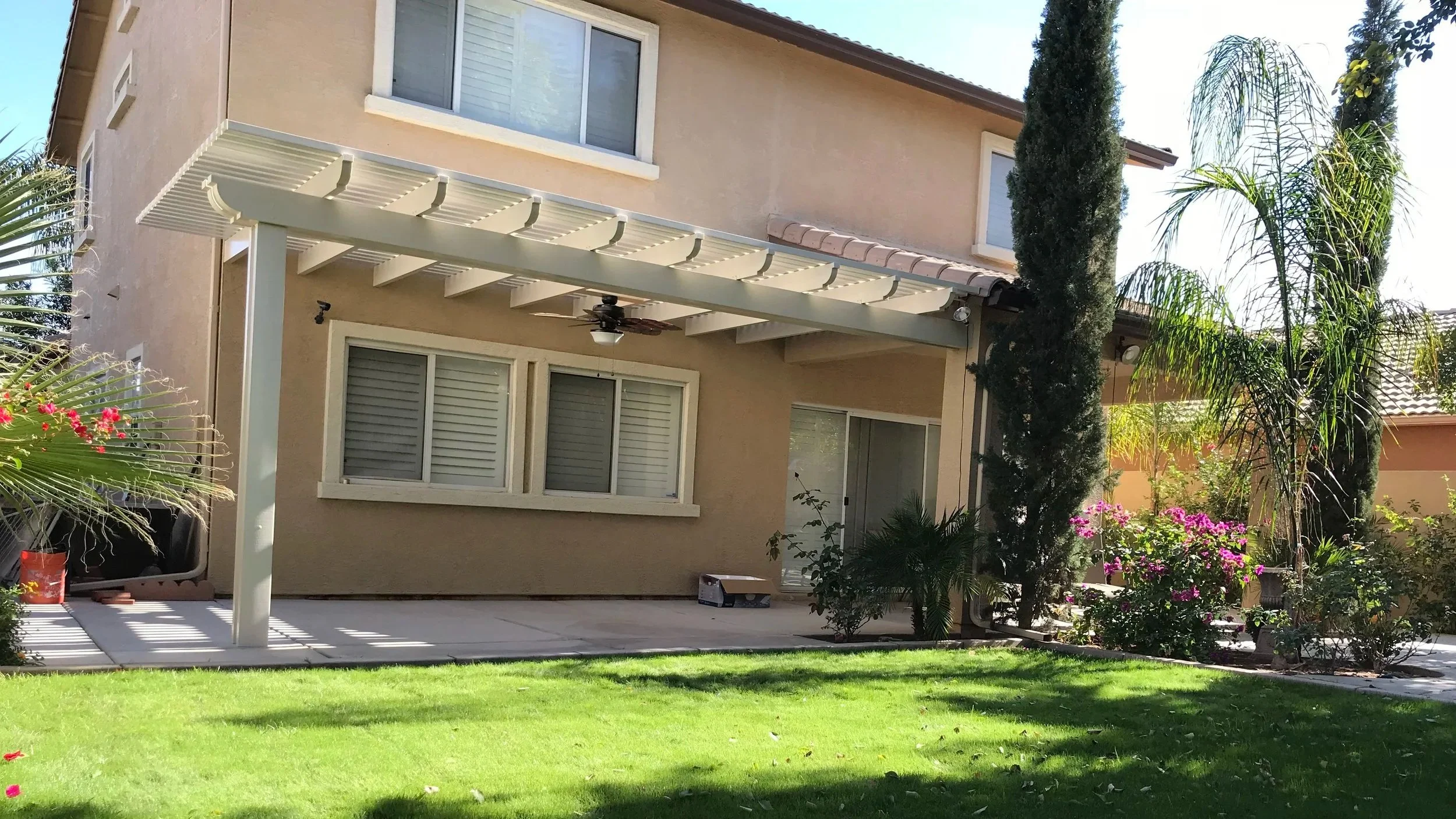 Backyard of a beige house with a covered patio, green grass, and various plants and trees including palms and a tall, narrow tree.