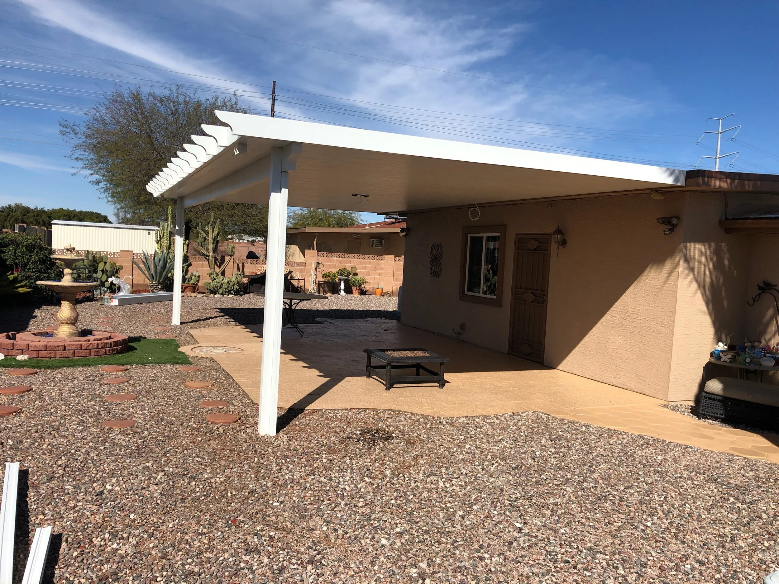 A backyard patio area with a beige stucco house, a covered patio with a white ceiling and wooden flooring, desert landscaping with cactus plants, a small fountain, and outdoor furniture under a clear blue sky.