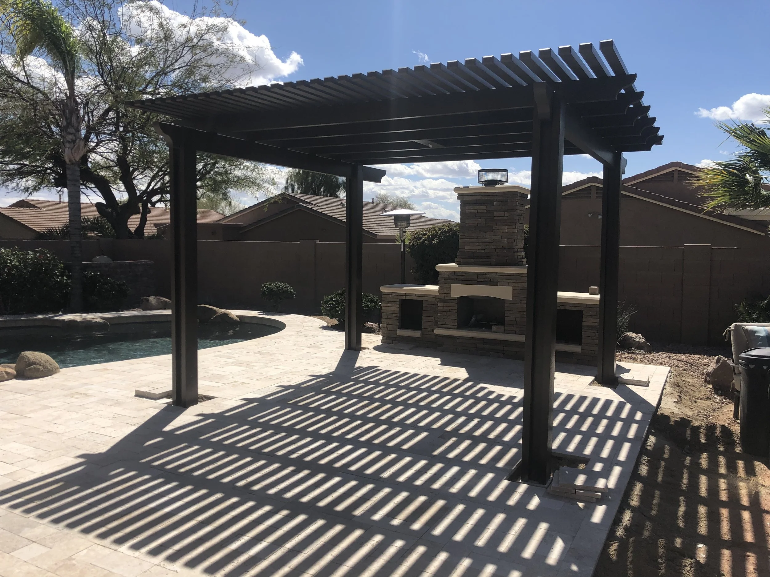 Patio area with a wooden pergola casting striped shadows, a brick outdoor fireplace, a swimming pool, and surrounding shrubbery in a residential backyard.