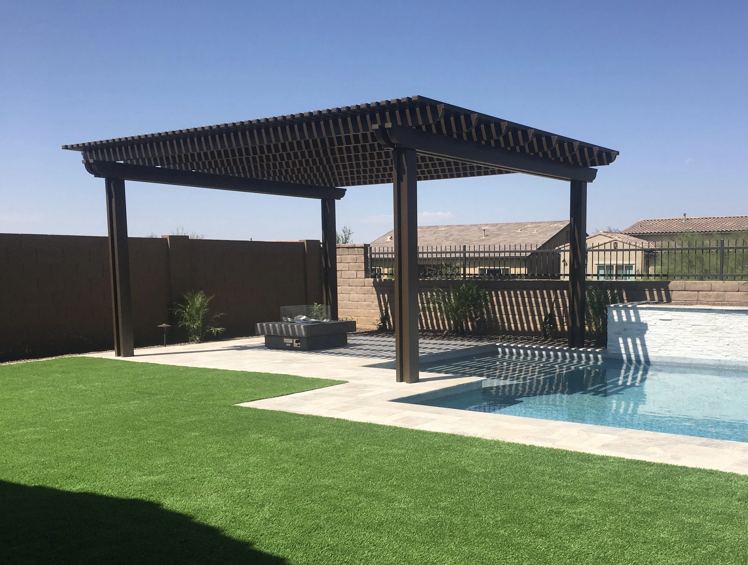 Backyard with a swimming pool, a pergola with a slatted roof, a small water fountain, and a well-maintained green lawn bordered by a brown wooden fence and a brick wall, under a clear blue sky.