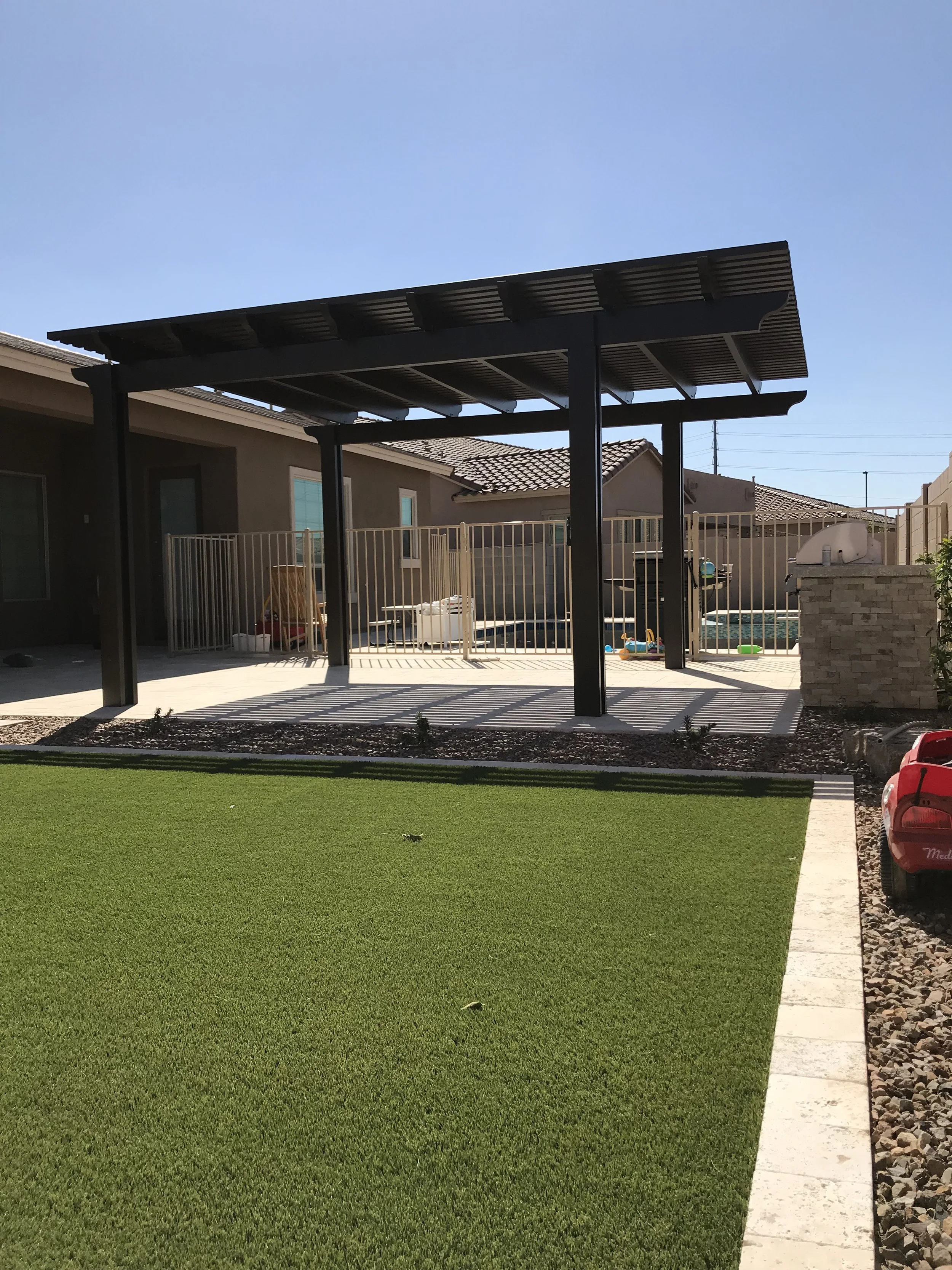 Backyard with a covered patio, artificial grass, and a small fence around a pool area. There is a red toy car on the ground to the right.