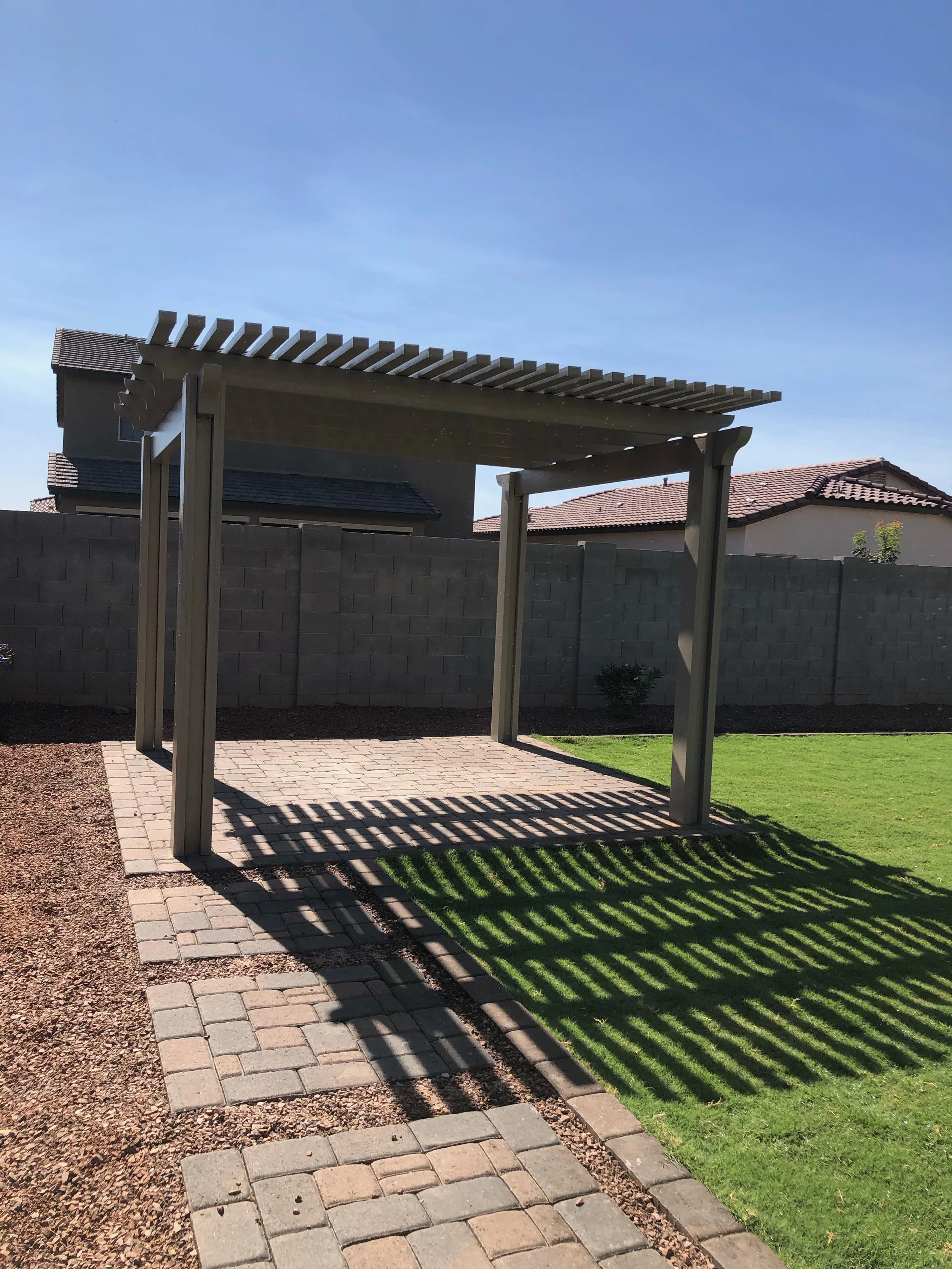 A wooden pergola in a backyard, casting striped shadows on the ground, with grass on one side and a brick pathway on the other.