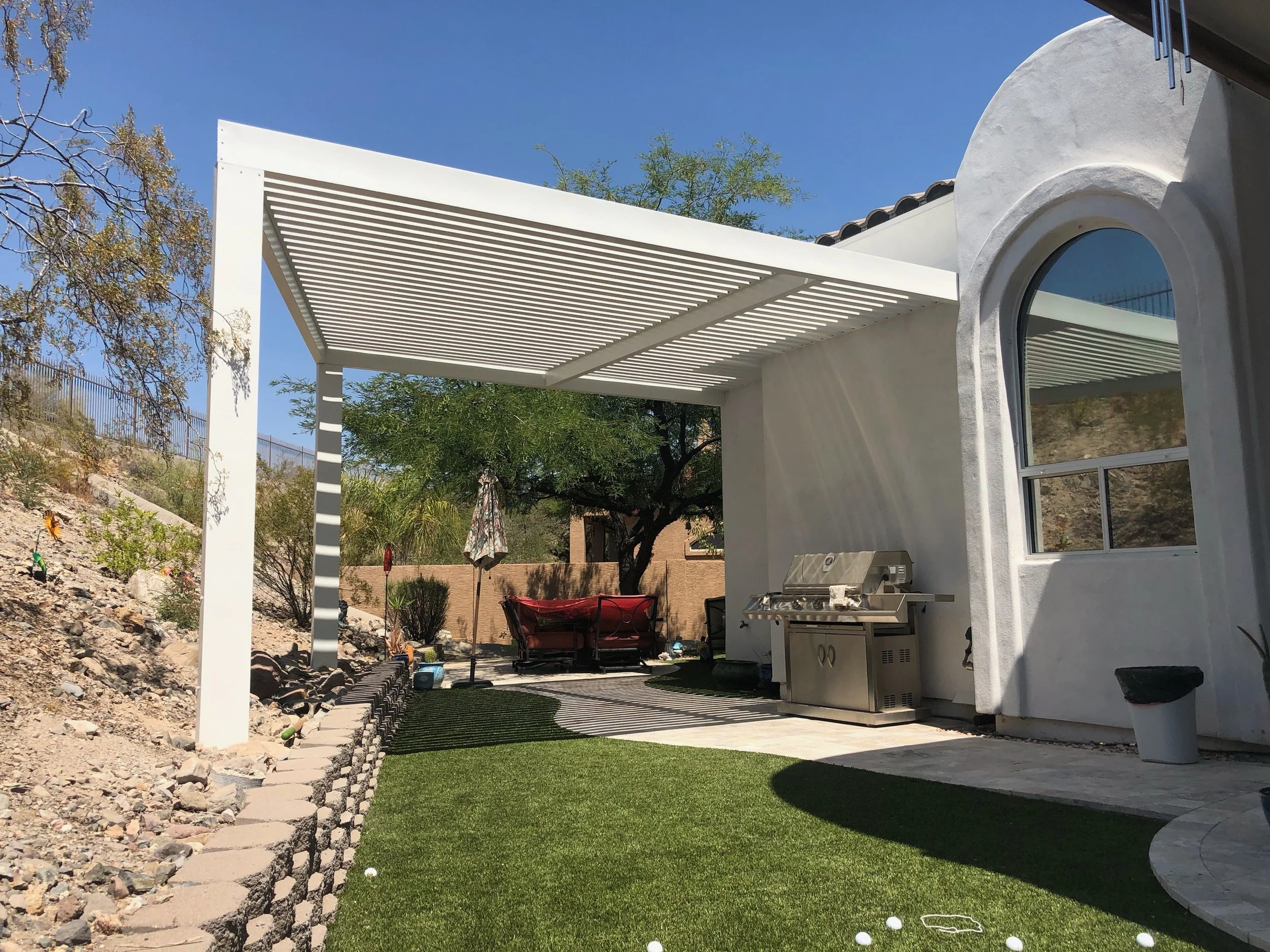 Backyard patio area with a white pergola, a grill, an outdoor seating area with a red couch, a white house wall with a window, and artificial grass.