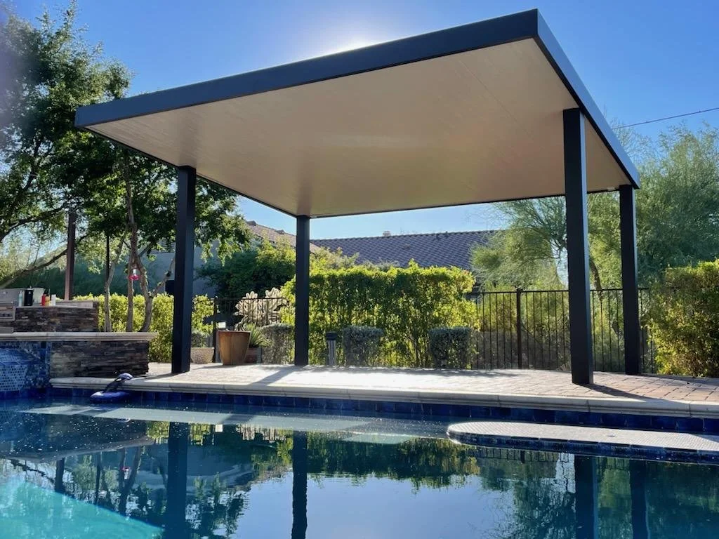 Poolside patio with a modern canopy structure, surrounded by lush greenery and a black fence, on a sunny day.
