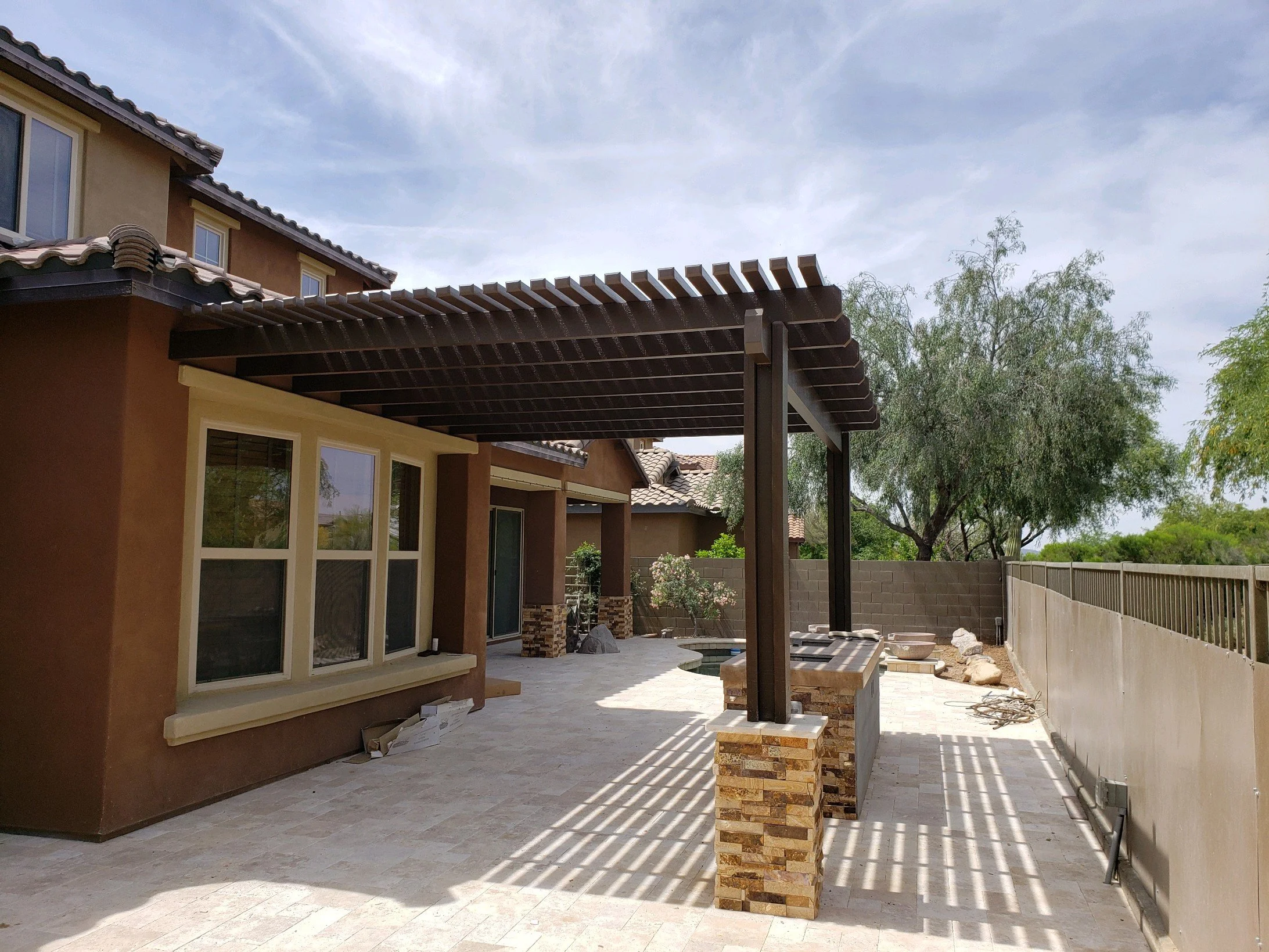 A modern house patio with a wooden pergola, stone bar counter, and a concrete block wall, with trees in the background under a partly cloudy sky.