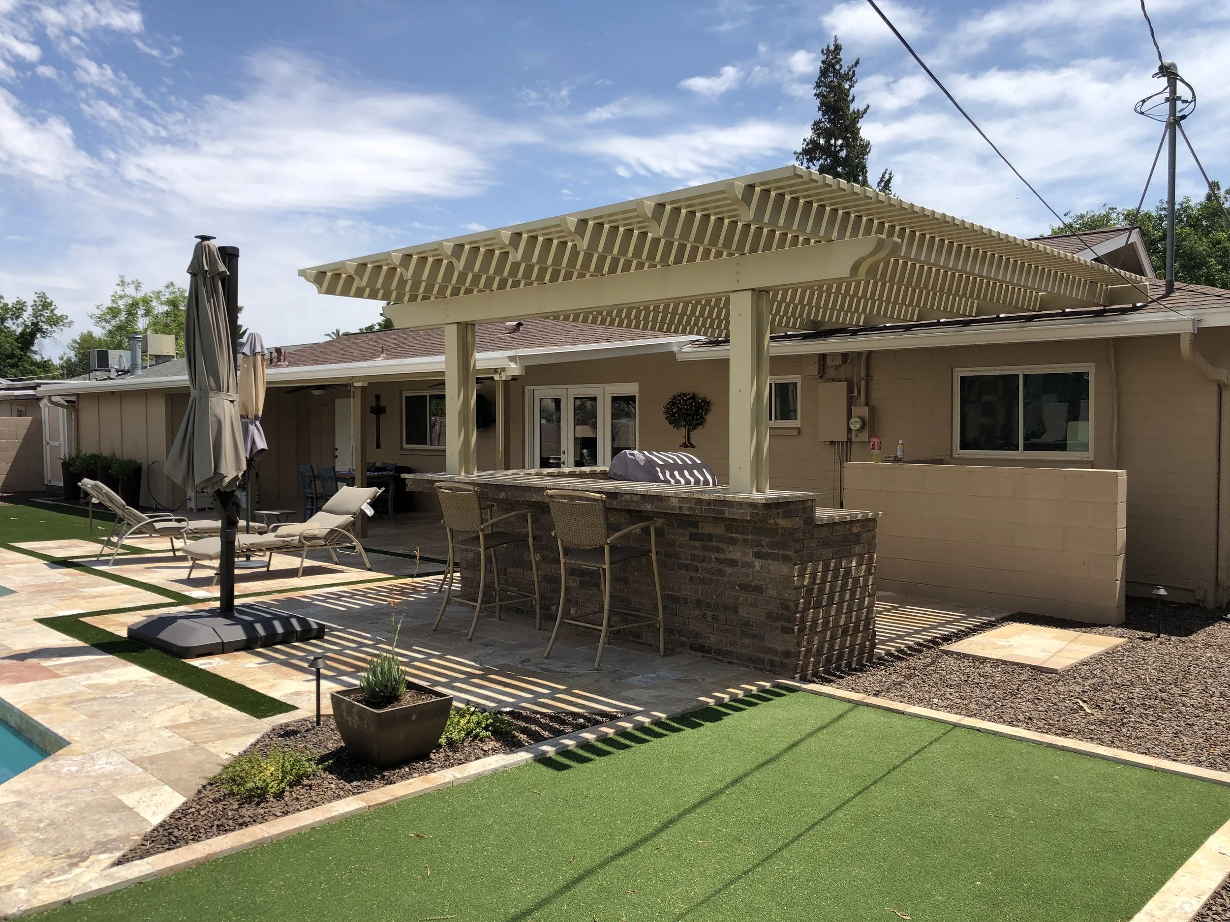 Residential backyard patio with outdoor furniture, pergola, and artificial grass area under a blue sky.