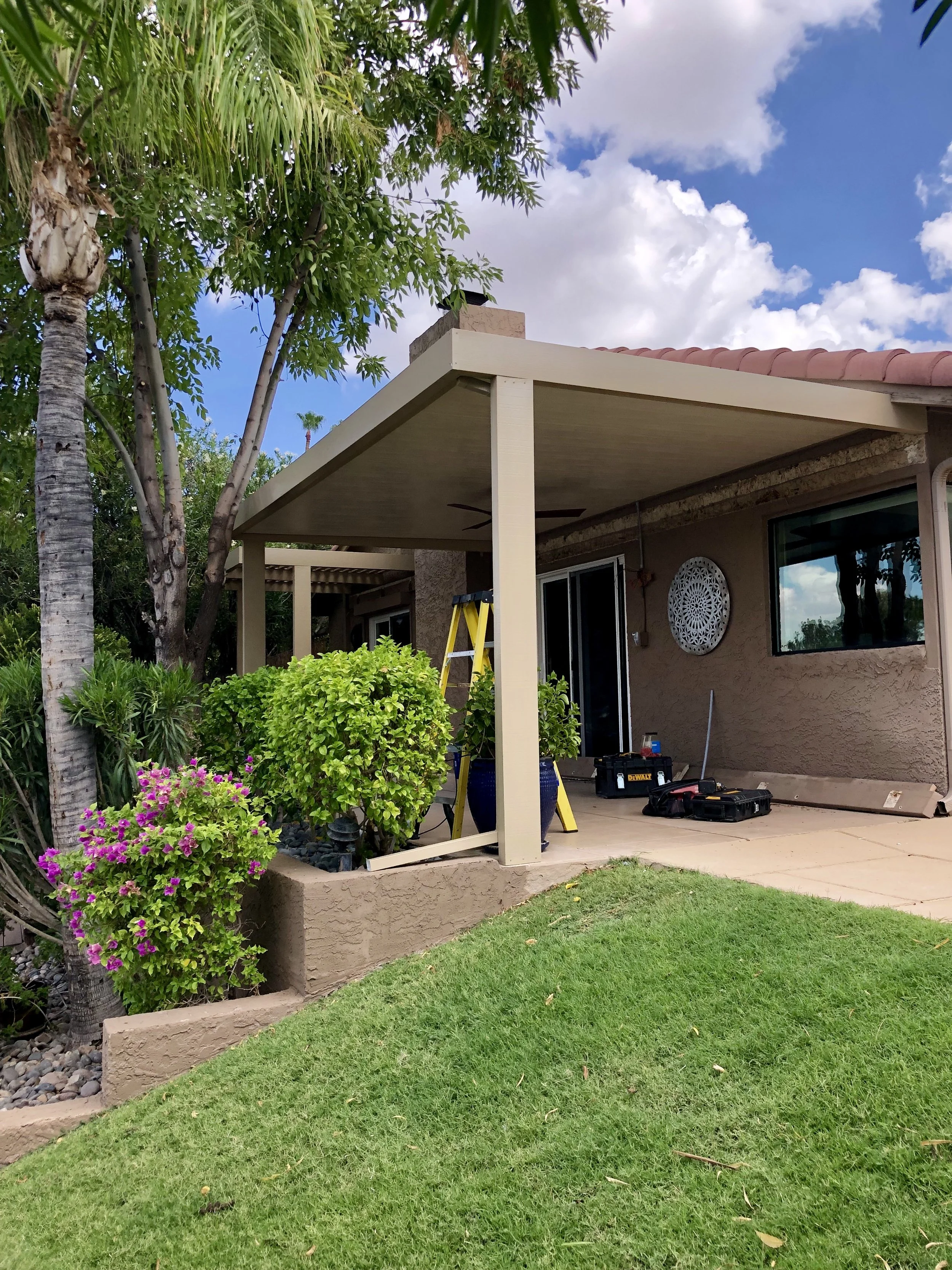 Backyard patio with construction tools, plant pots, and a ladder, part of a house with beige stucco walls, sliding glass doors, plants, trees, and a grassy lawn.