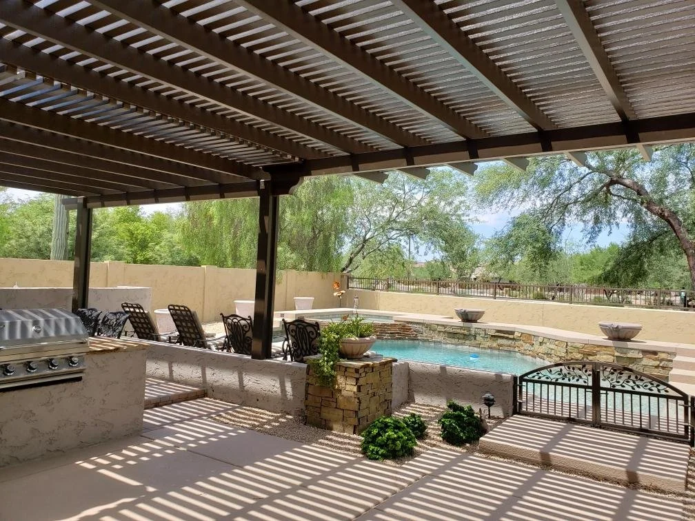 Covered patio with outdoor seating, grill, and view of a backyard pool surrounded by trees and a beige wall.