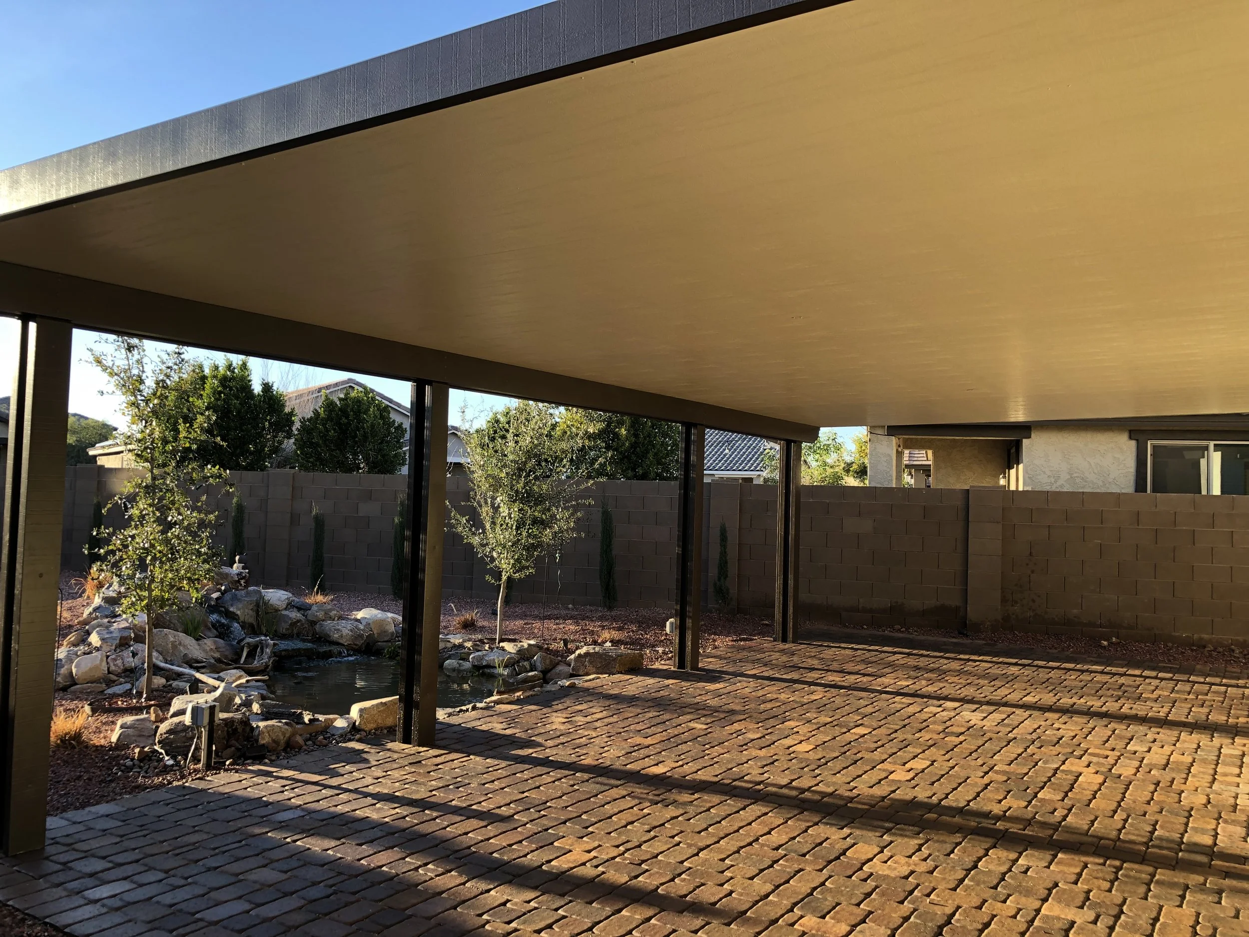 View of a covered outdoor patio with brick flooring, a small pond with rocks, and trees in the backyard, enclosed by a brick wall.
