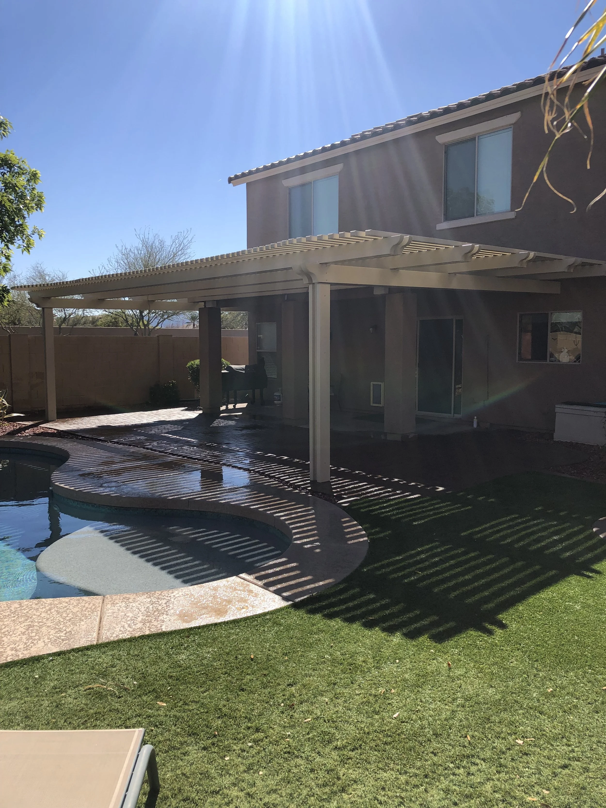 Backyard with a swimming pool, covered patio area with outdoor furniture, and a two-story house with windows, under a bright sunny sky.