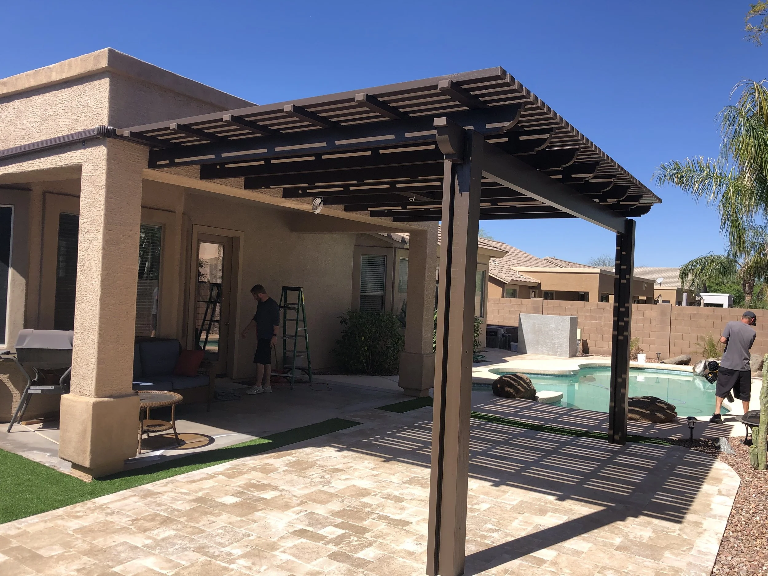 Backyard with covered patio, swimming pool, and two men working outside on a sunny day.