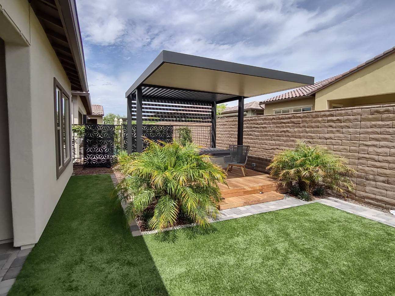 Backyard with artificial grass, a wooden patio with a seating area, plants, a privacy fence, and a covered pergola under a cloudy sky.