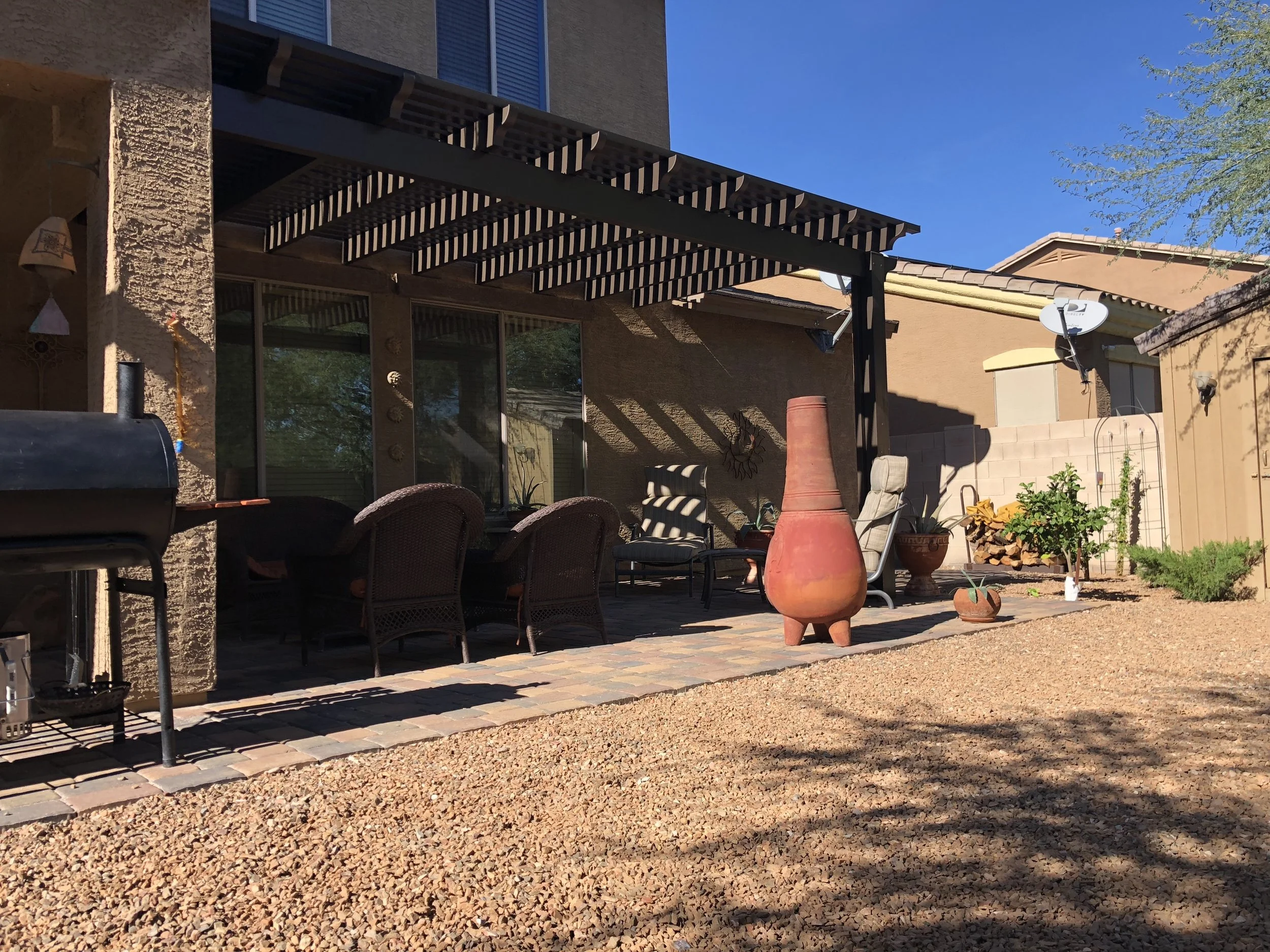 Backyard patio with chairs, potted plants, and decorative pottery, partly shaded by a pergola, with a gravel ground and neighboring houses in the background.