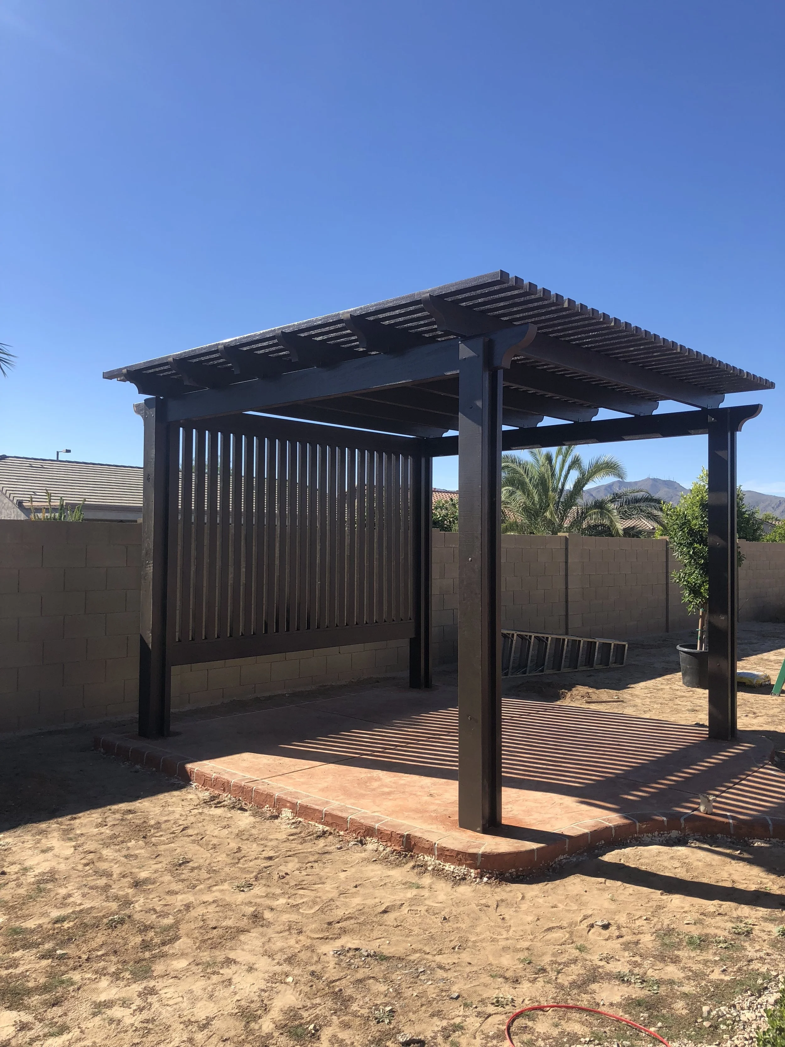 A black wooden pergola with a slatted roof and open sides over a brick patio, in a backyard with a dirt ground, a brick wall, and palm trees under a clear blue sky.