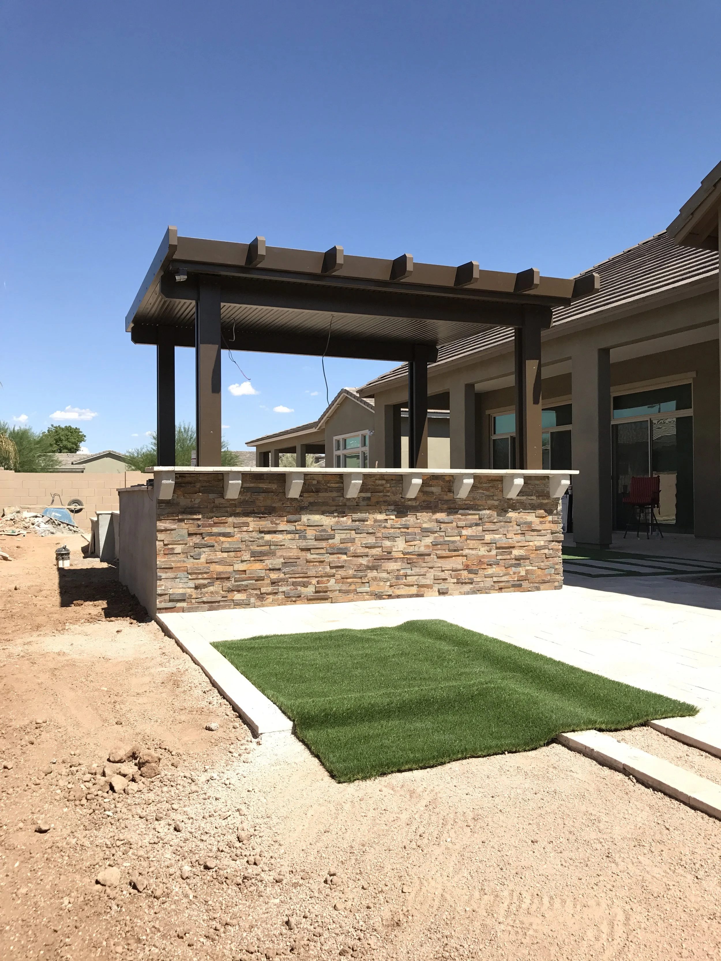 Backyard with outdoor patio area, stone wall, pergola, artificial grass patch, and part of house with glass sliding doors
