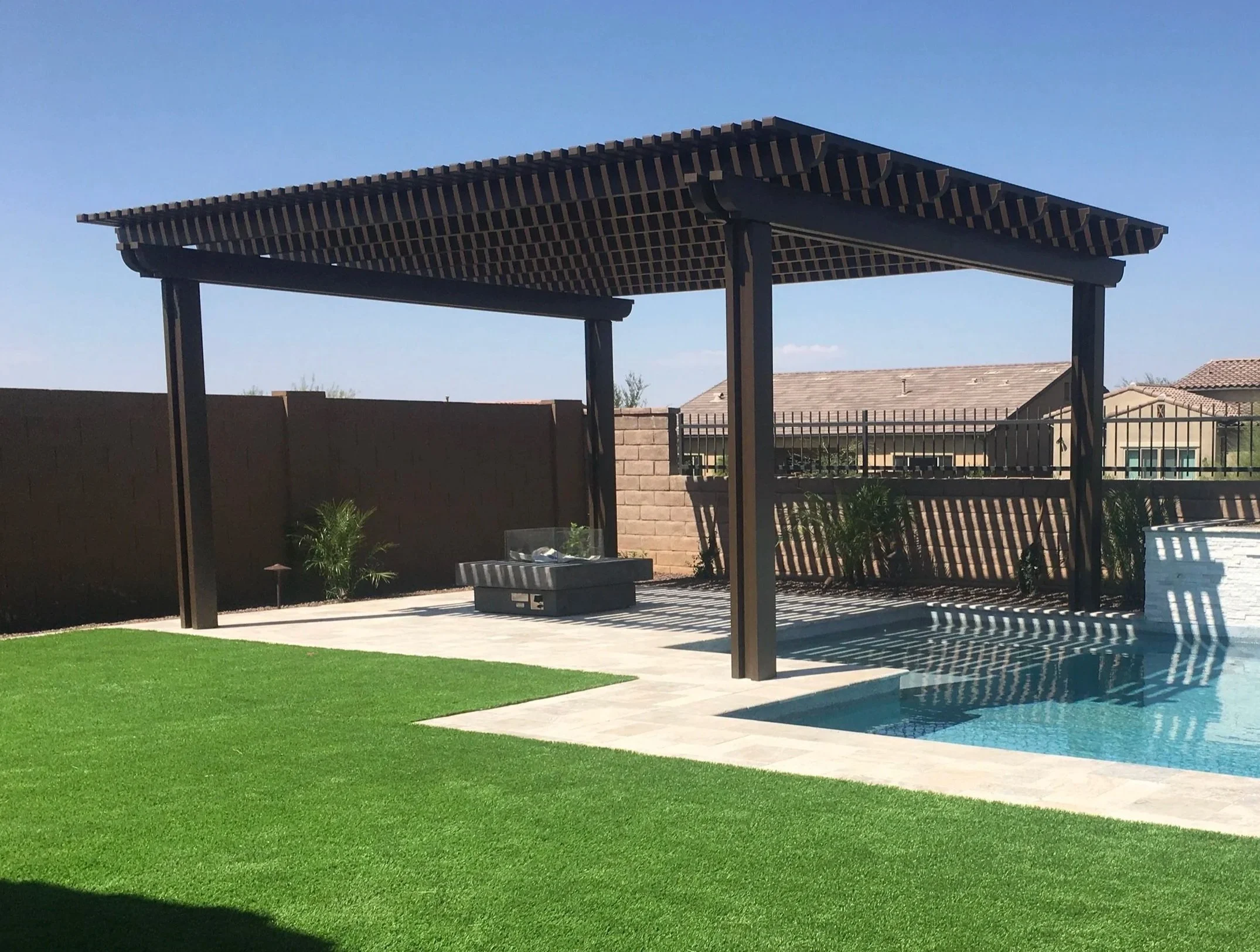 Backyard with swimming pool, green lawn, and a wooden pergola with lattice roof, surrounded by privacy fence and small plants.