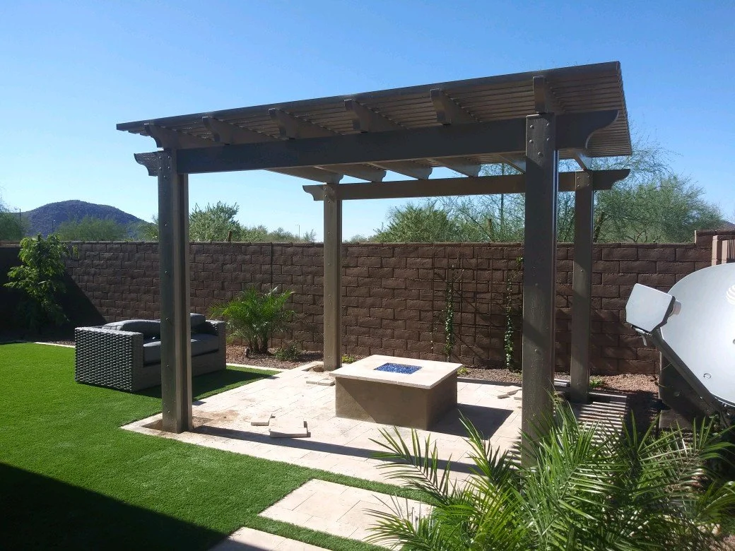 Backyard patio with a wooden pergola, a fire pit, a gray outdoor sofa, a satellite dish, and desert landscape with mountains in the background.