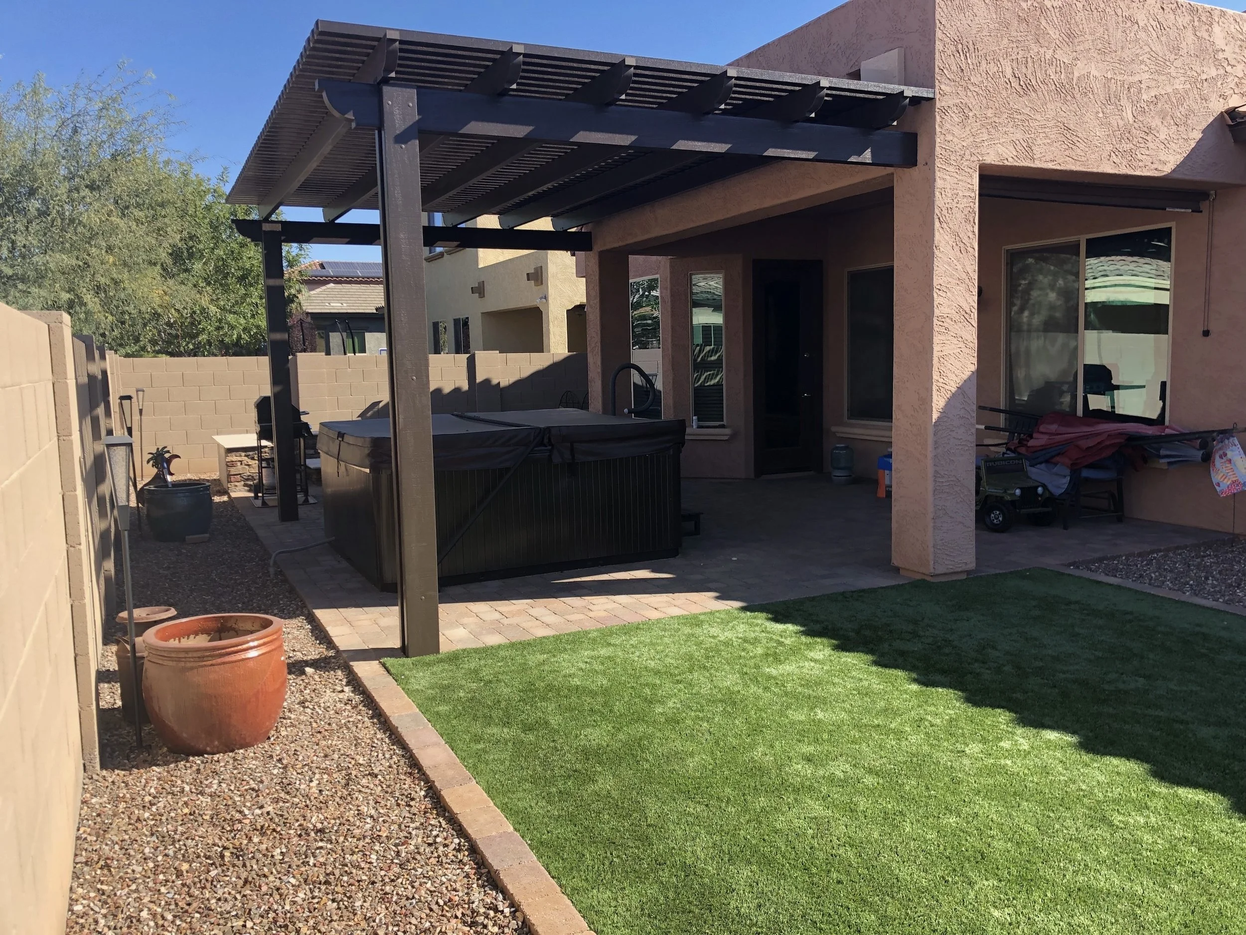 Backyard patio with artificial grass, a hot tub under a pergola, and a backyard with potted plants, gravel, and outdoor furniture.