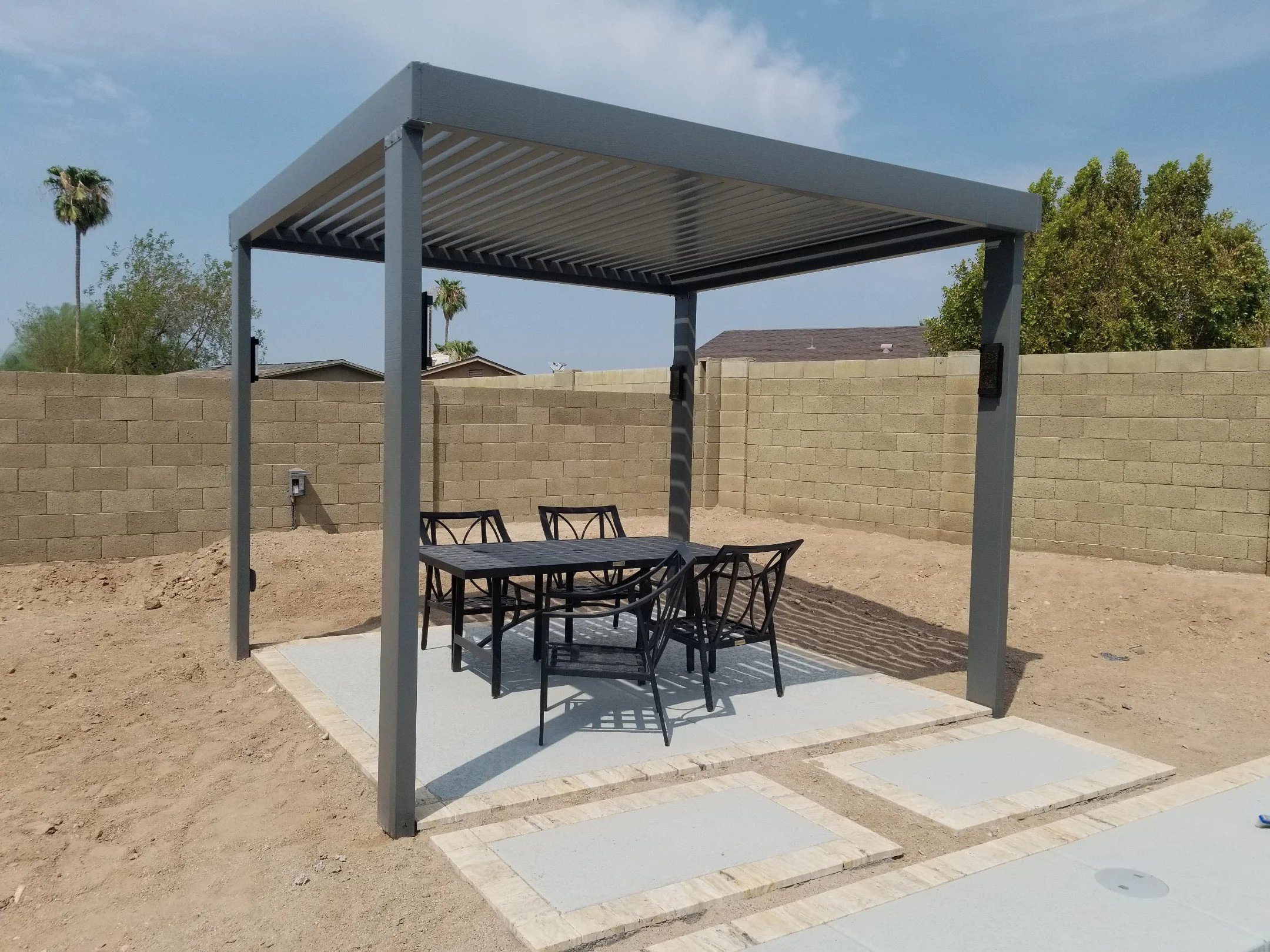 Outdoor covered patio with a table and six chairs on a concrete slab, surrounded by a dirt yard and a brick wall, with trees and houses in the background.