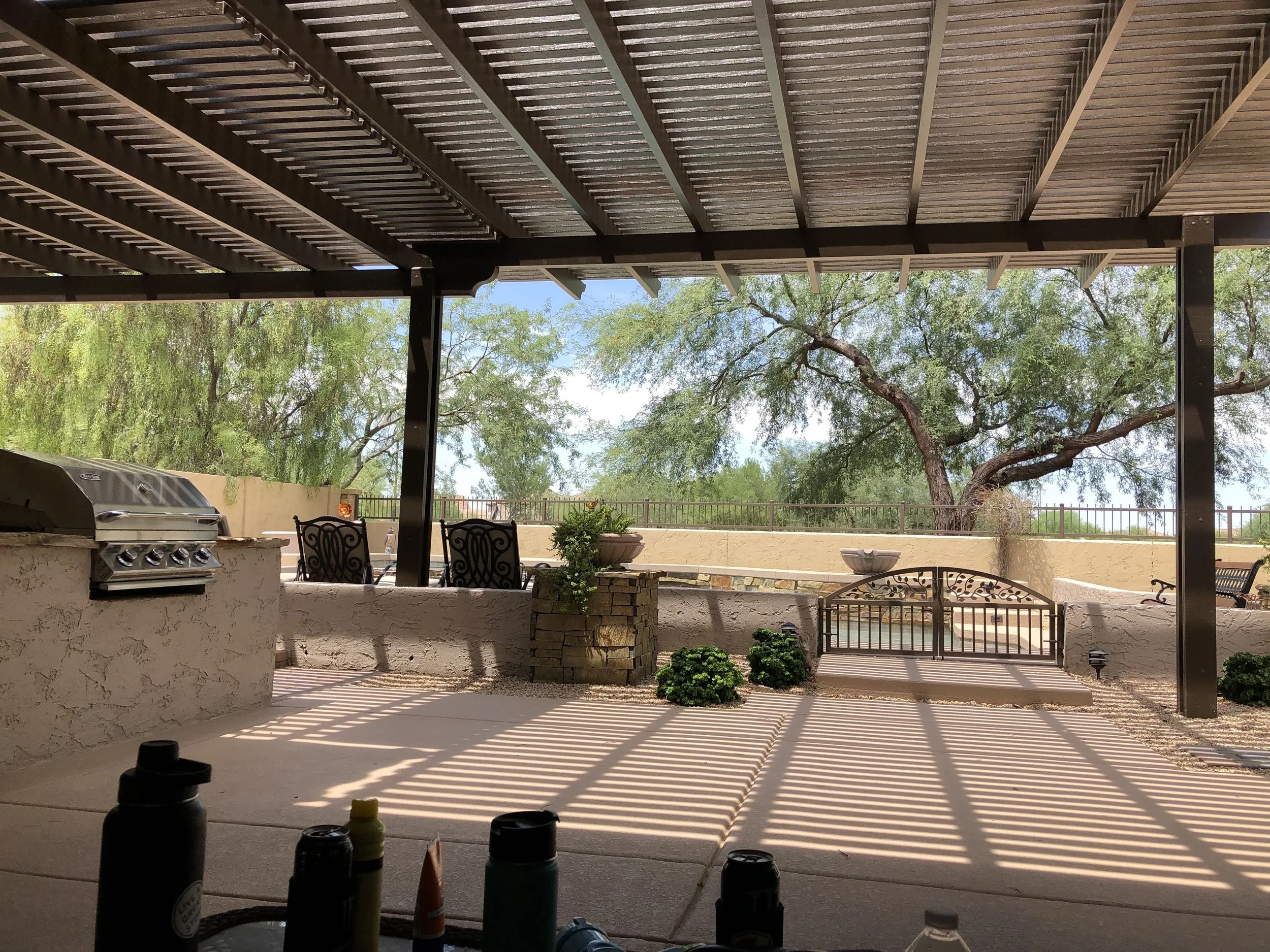 Back patio area shaded by a roof with a view of trees and a landscaped yard with small shrubs and a fire pit.