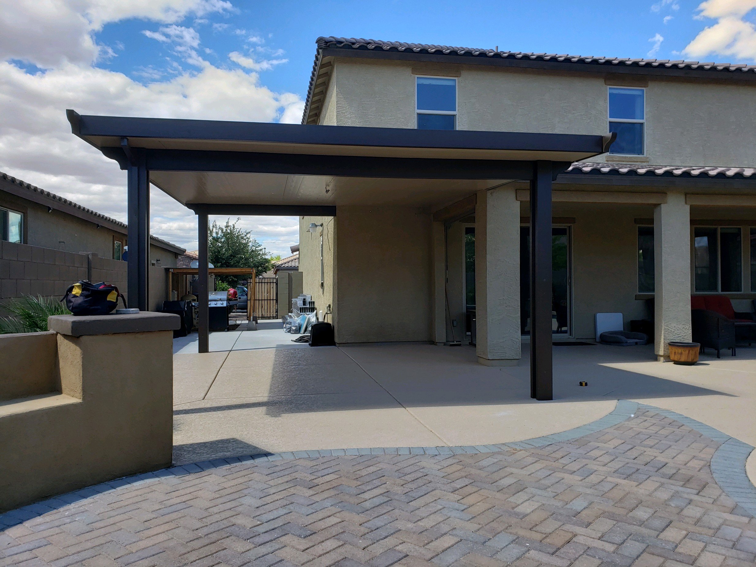 Backyard patio area with a covered section, some outdoor furniture, and a house with beige walls and a tiled roof. There are items like bags, bottles, and a grill near the covered area, and a cloudy sky above.