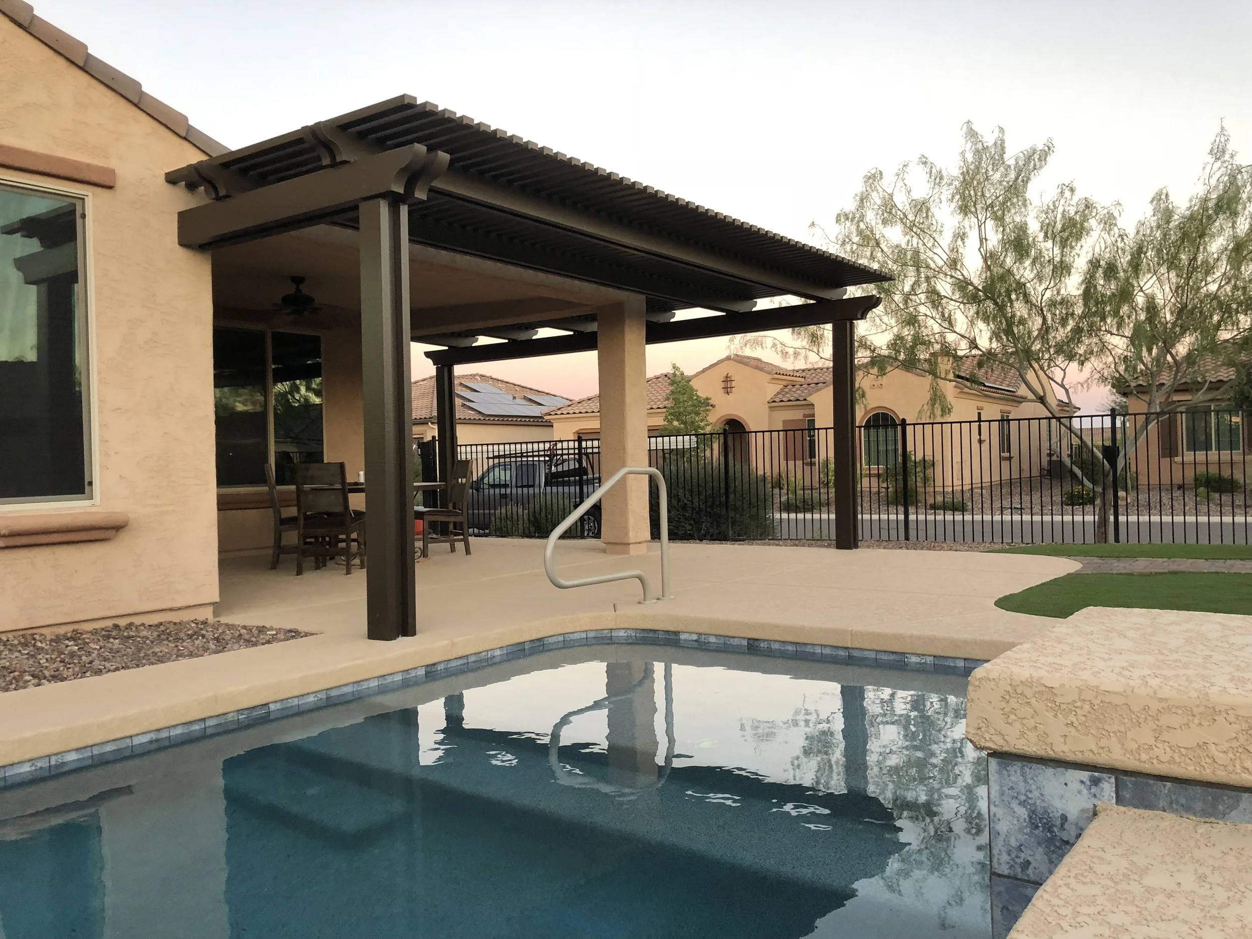 Residential backyard with a swimming pool, covered patio area with a dining table and chairs, a black metal fence, trees, and neighboring houses in the background.