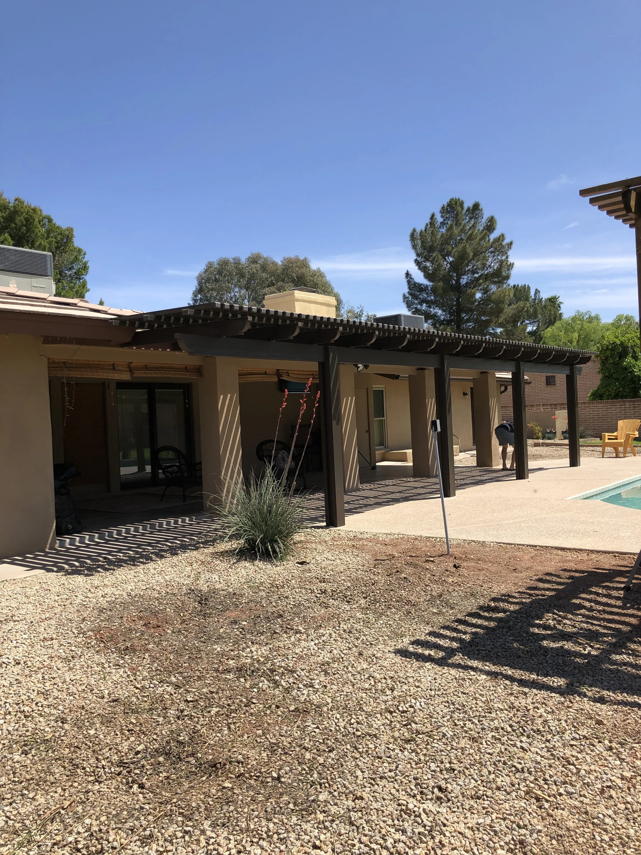 Rear view of a house with a covered patio, outdoor furniture, a swimming pool, and a few trees in the background, under clear blue skies.