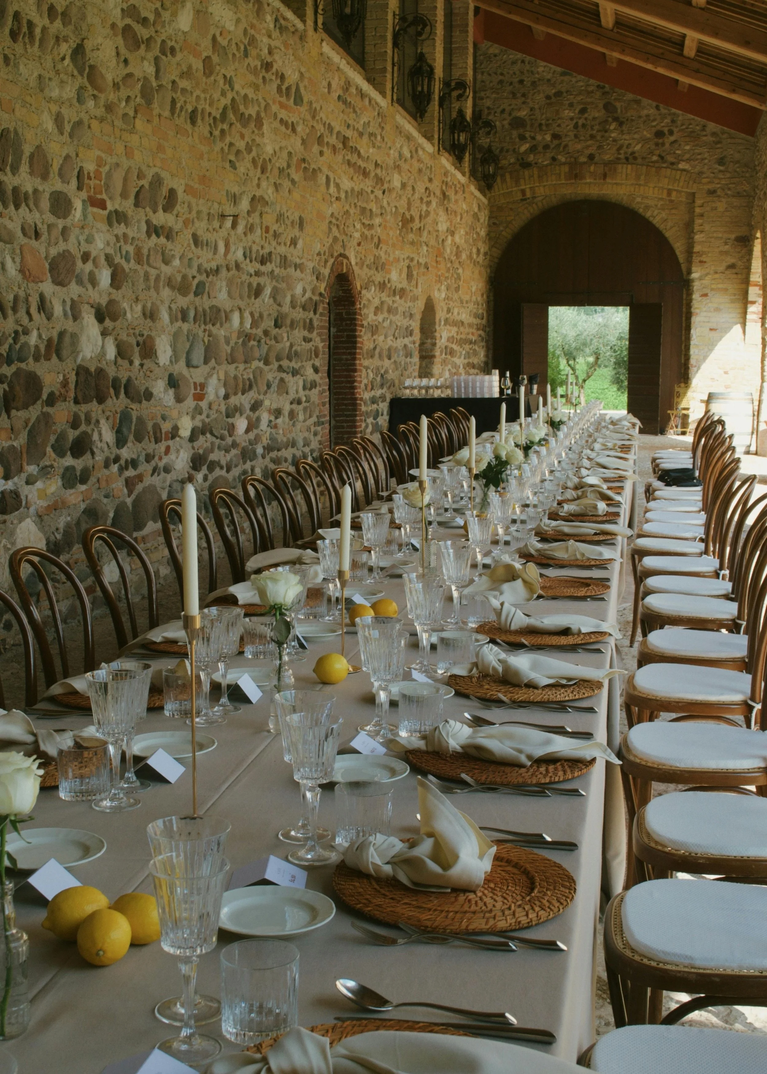 A long table set for a formal meal in a rustic stone barn, with white tablecloths, candles, glassware, and lemons, arranged for an event or celebration.