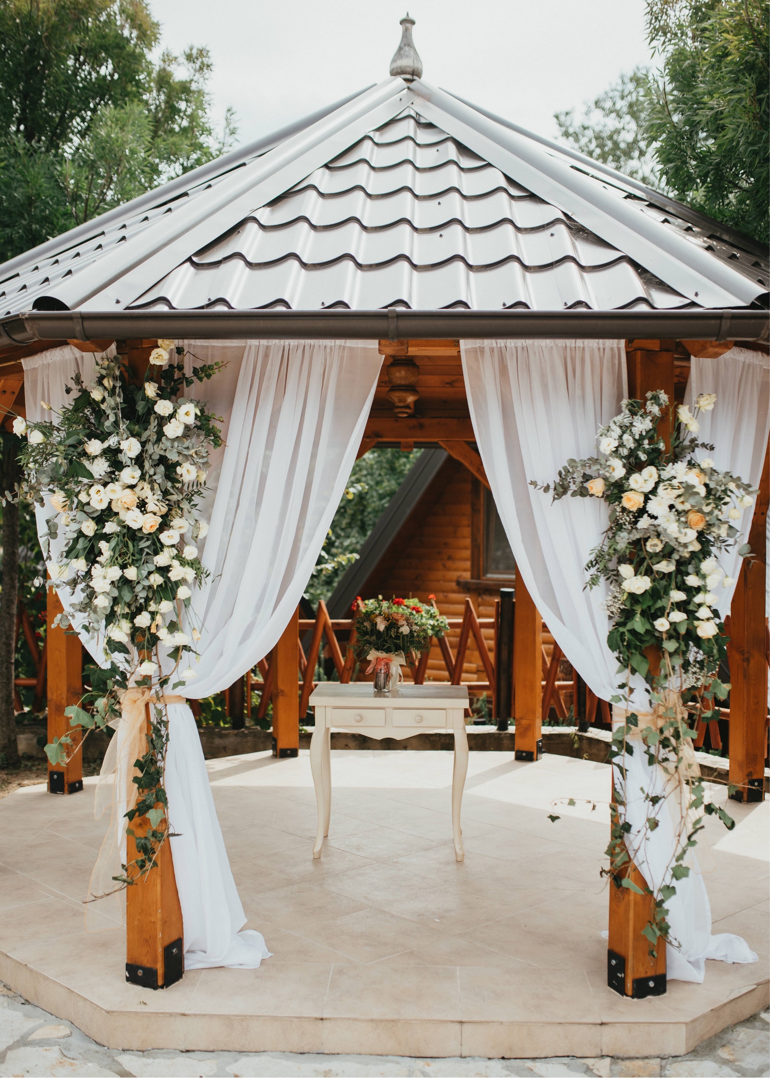 Decorative outdoor wedding gazebo with white drapes and floral arrangements, set on a tiled platform.