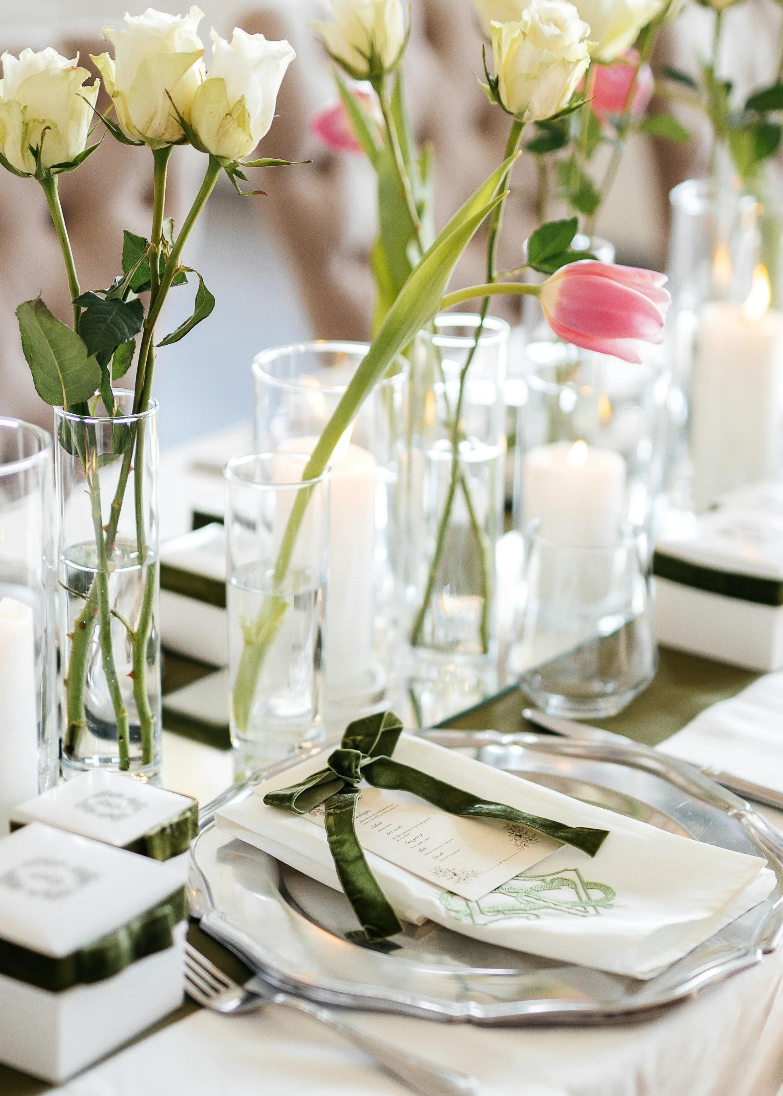 Elegant table setting with white roses and pink tulips in glass vases, candles, and place setting with wrapped utensils and a menu card on a white cloth.
