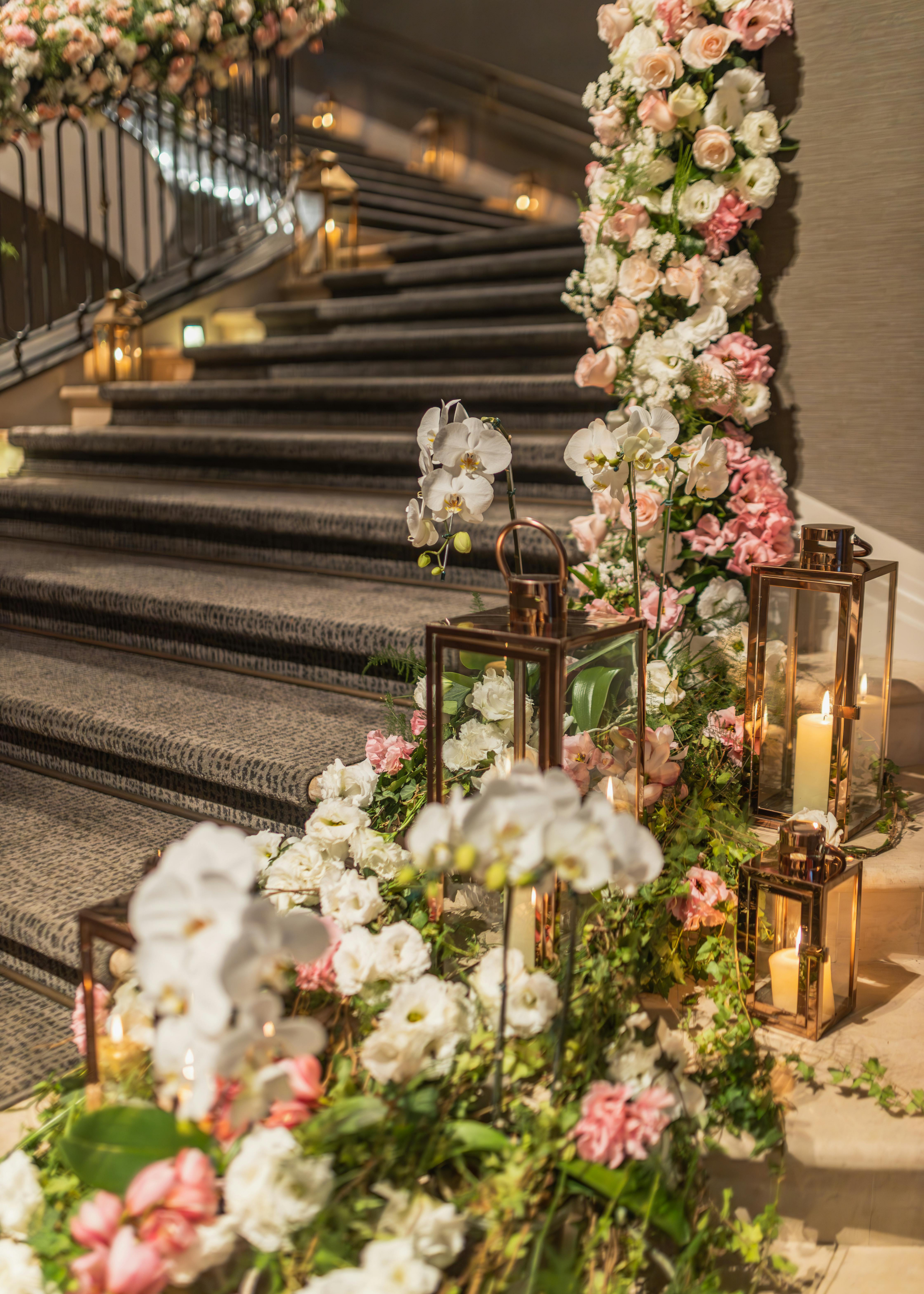 Decorated staircase with floral arrangements of white and pink flowers, bronze lanterns with candles, and soft lighting for an elegant event.