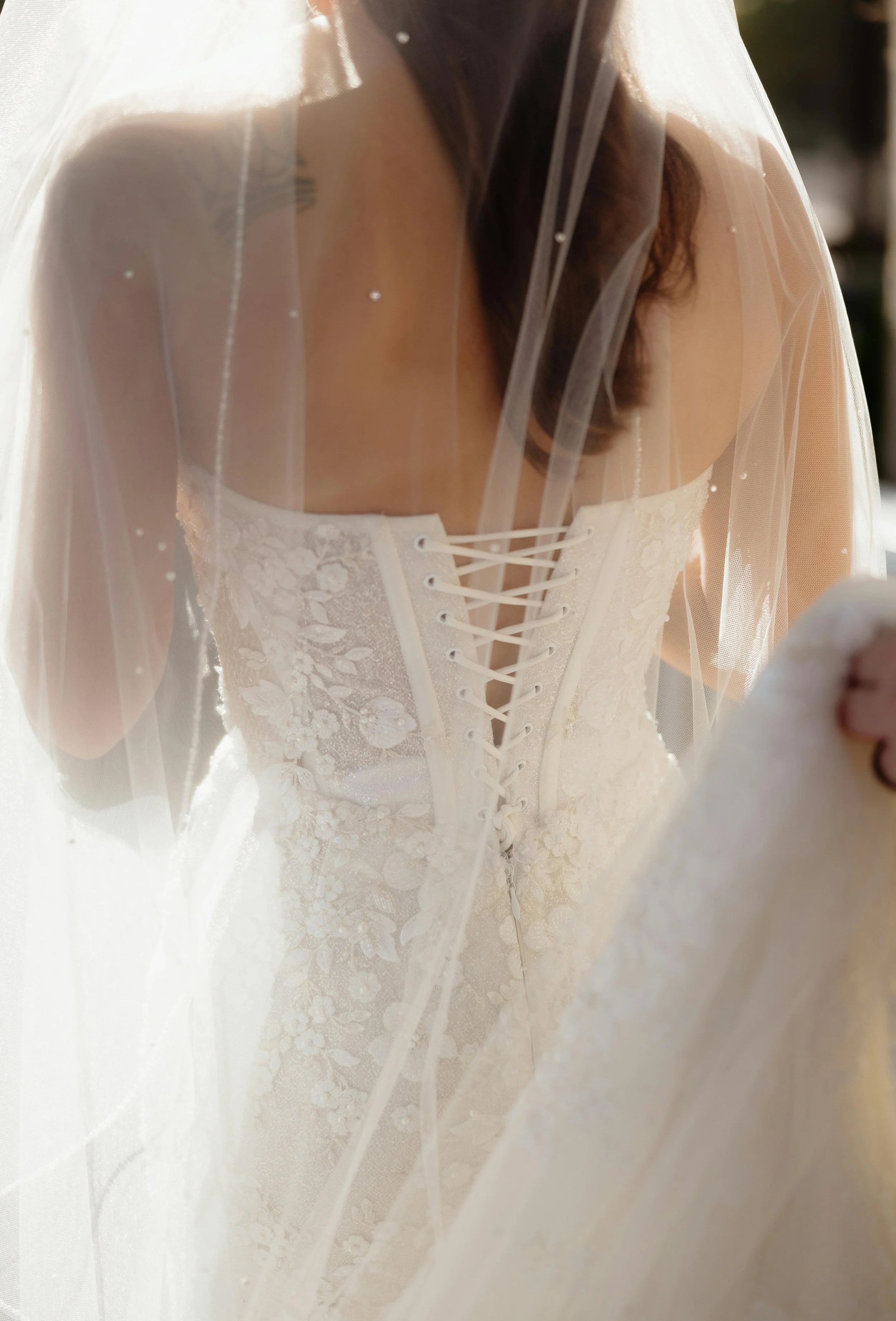 Close-up of a bride in a lace wedding dress under a veil, focusing on the front of the dress with a laced-up corset back.
