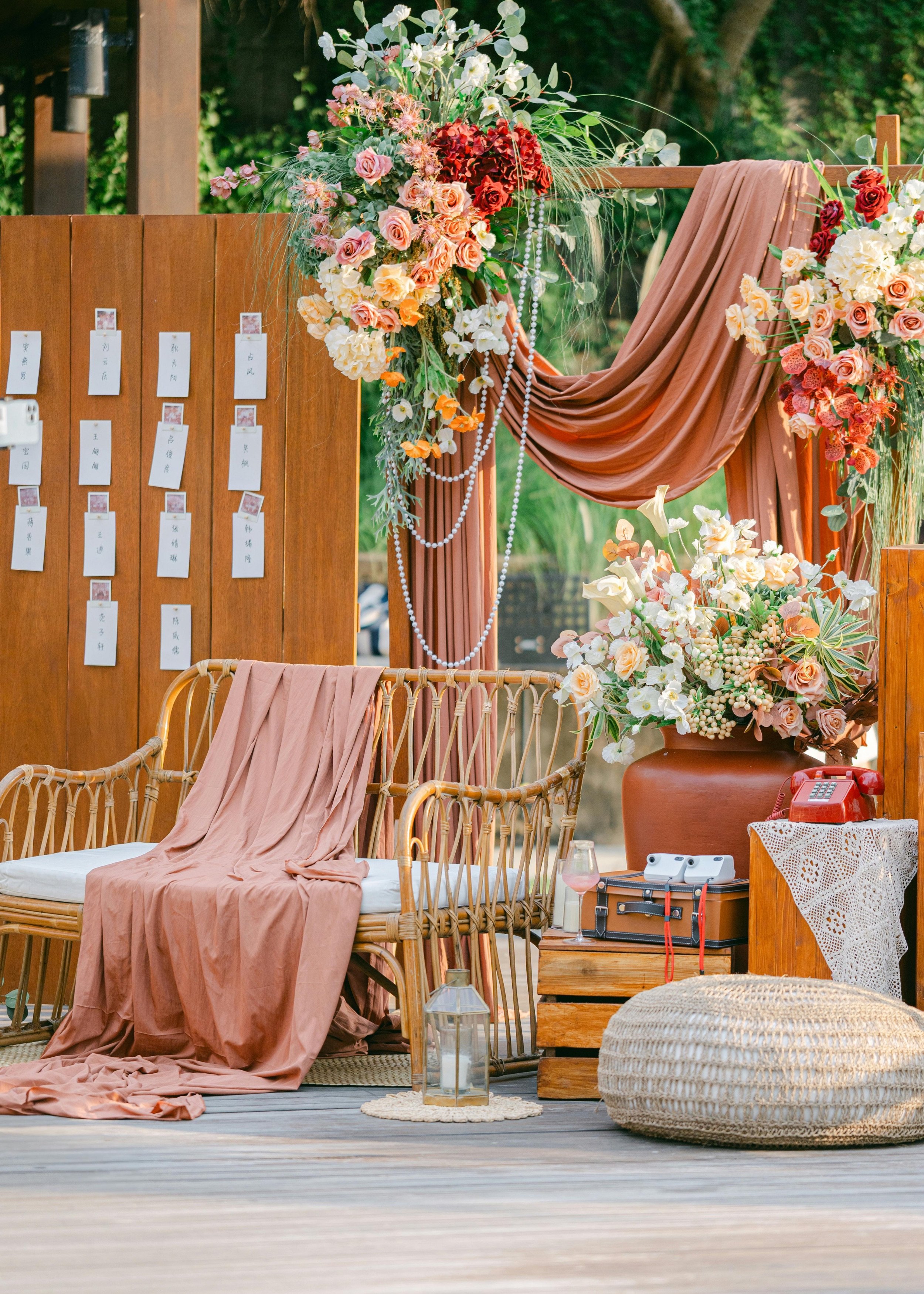 Decorative outdoor setting with pink and white flowers, draped fabric, a rattan bench with a pink cloth, a large flower arrangement in a terracotta vase, a red rotary phone, and a lantern on the wooden floor.