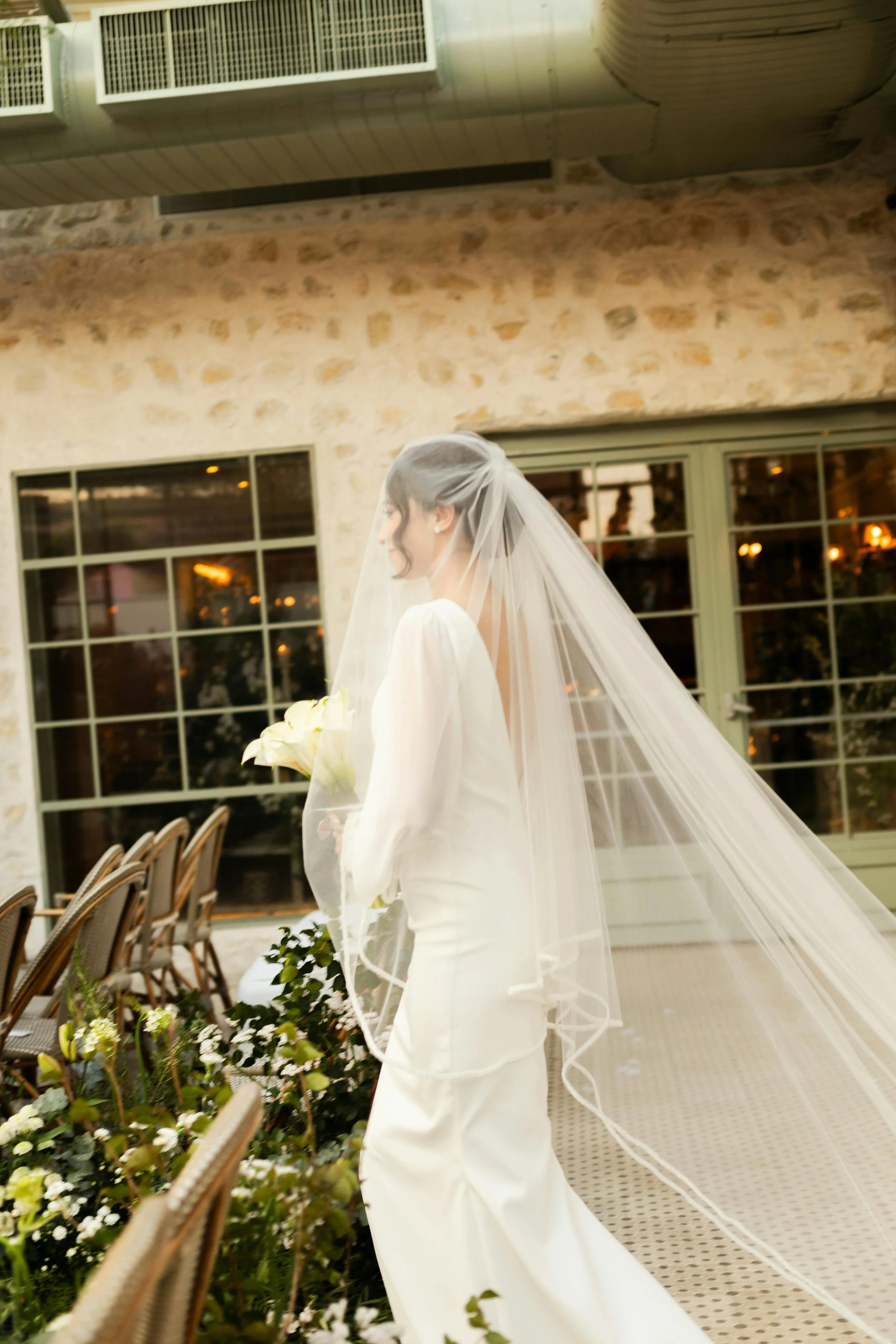 Bride in a white wedding dress with long veil holding a bouquet of flowers, standing near chairs and green plants in a rustic indoor setting with windows and stone walls.