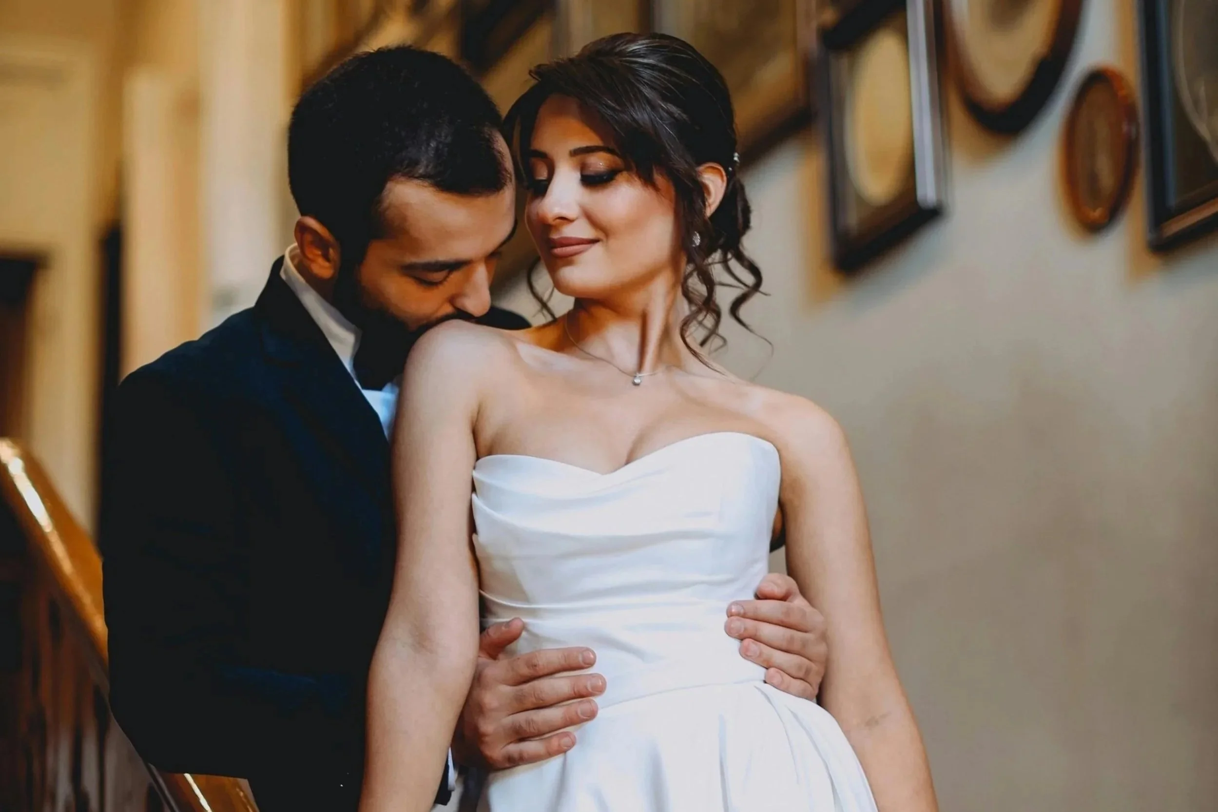 A newlywed couple in wedding attire sharing an intimate moment indoors, with the groom gently holding the bride's waist as she leans into him, smiling softly.