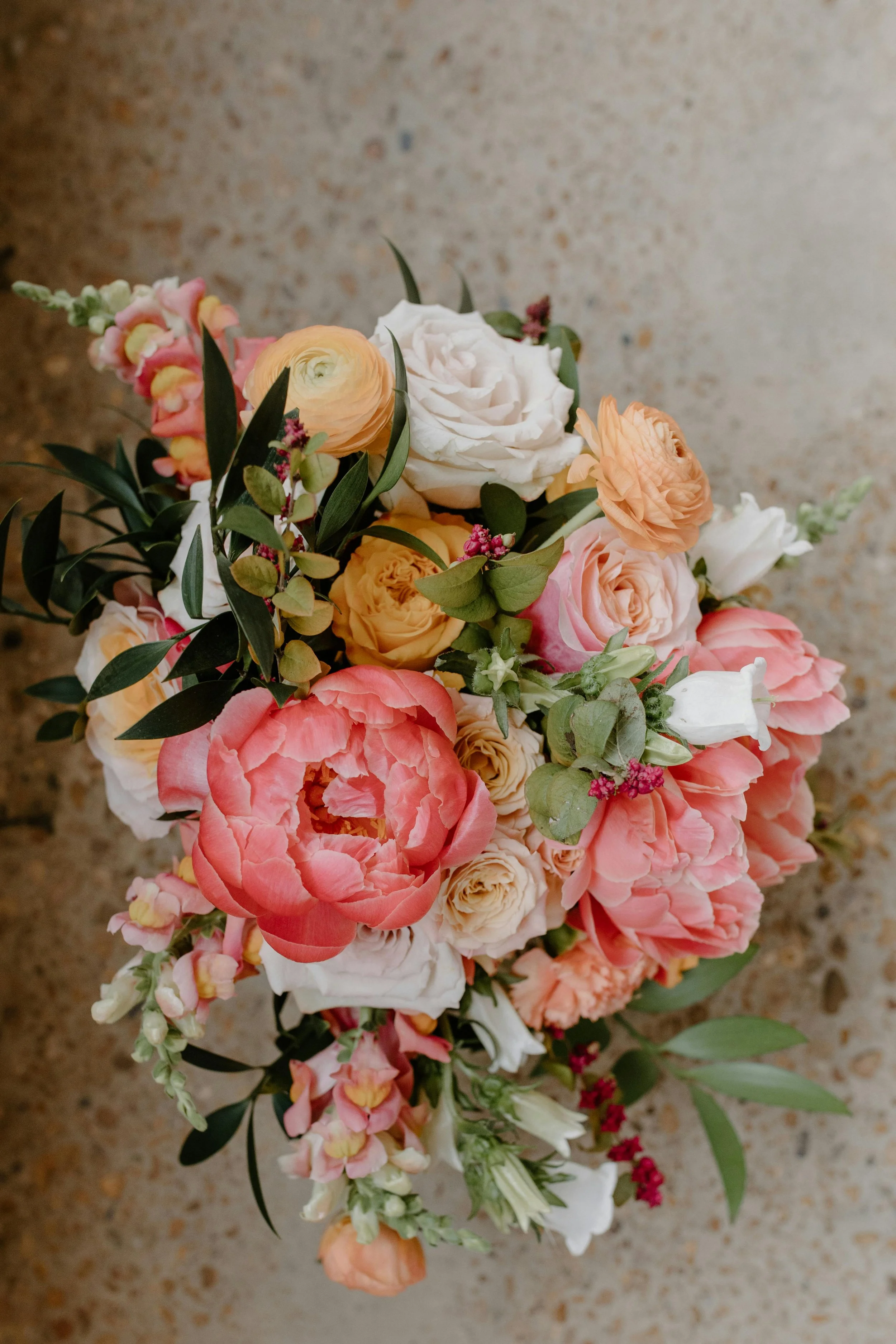 A bouquet of assorted flowers including white, peach, and pink roses, peach ranunculus, white calla lilies, and green foliage.
