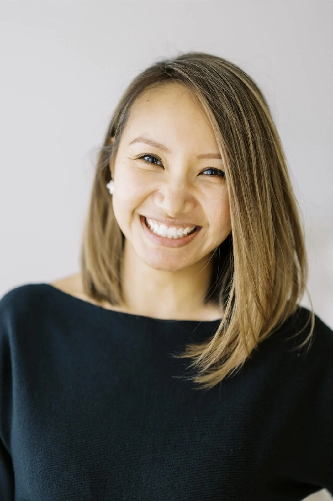 A smiling woman with shoulder-length brown hair, wearing a black top and small earrings, standing against a plain light-colored background.