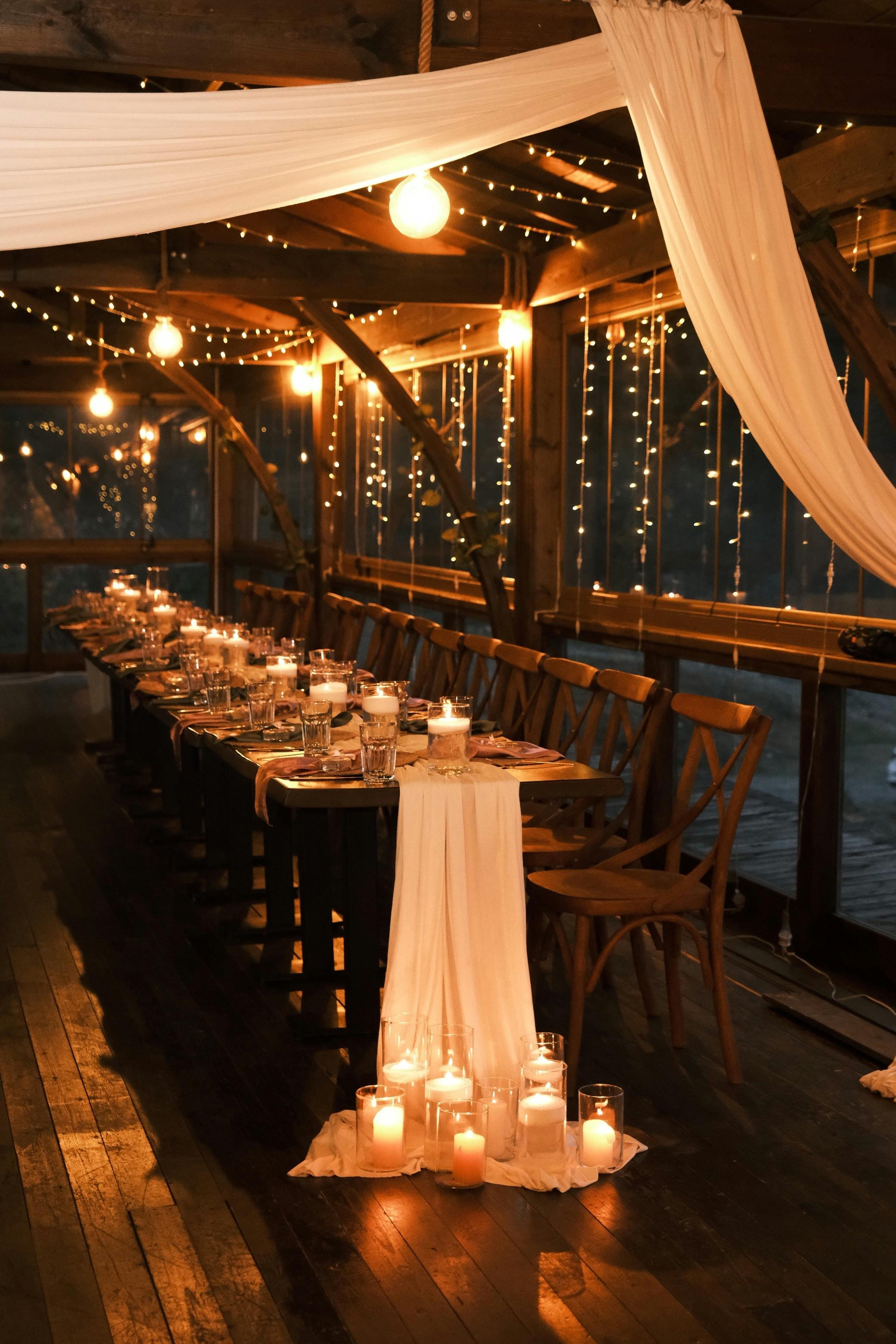 A warmly lit indoor dining table decorated with candles, glasses, and cloths, set for an event or celebration, with sheer fabric draped overhead and string lights hanging around.