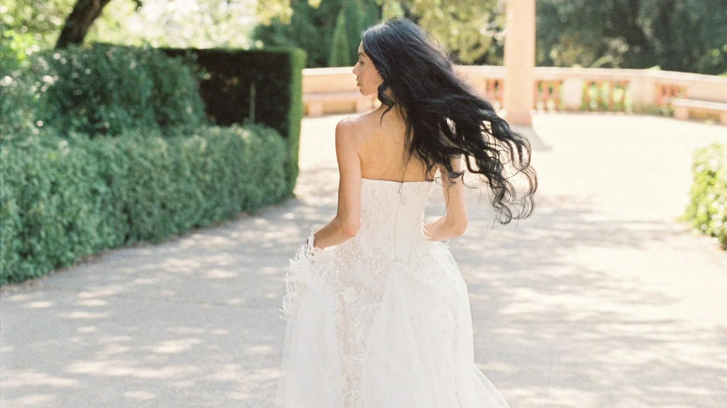 A woman in a white wedding dress walking outdoors with long black hair flowing behind her, surrounded by greenery and trees.