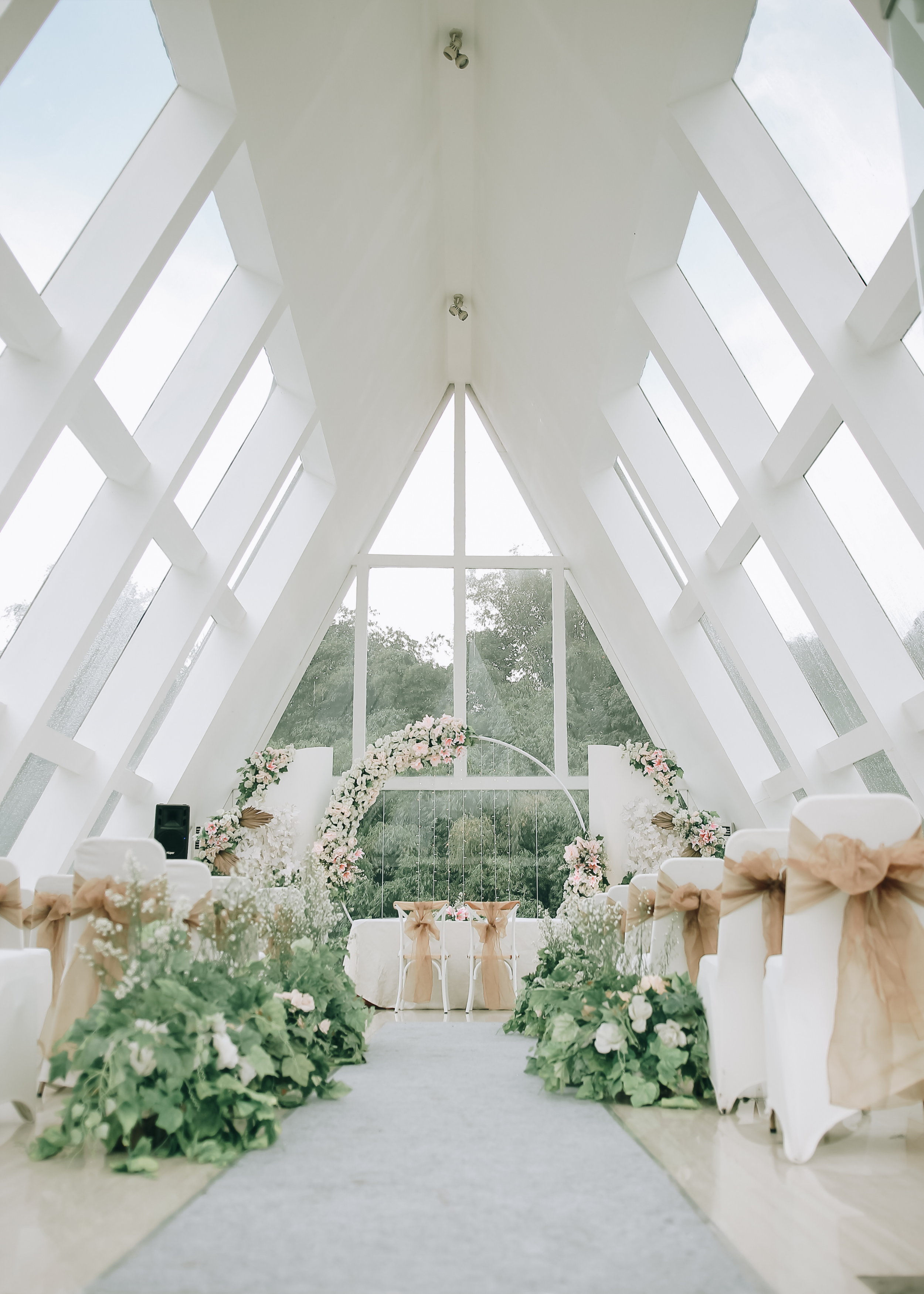Interior of a modern chapel with large glass windows, decorated for a wedding with floral arrangements and chairs with beige bows.