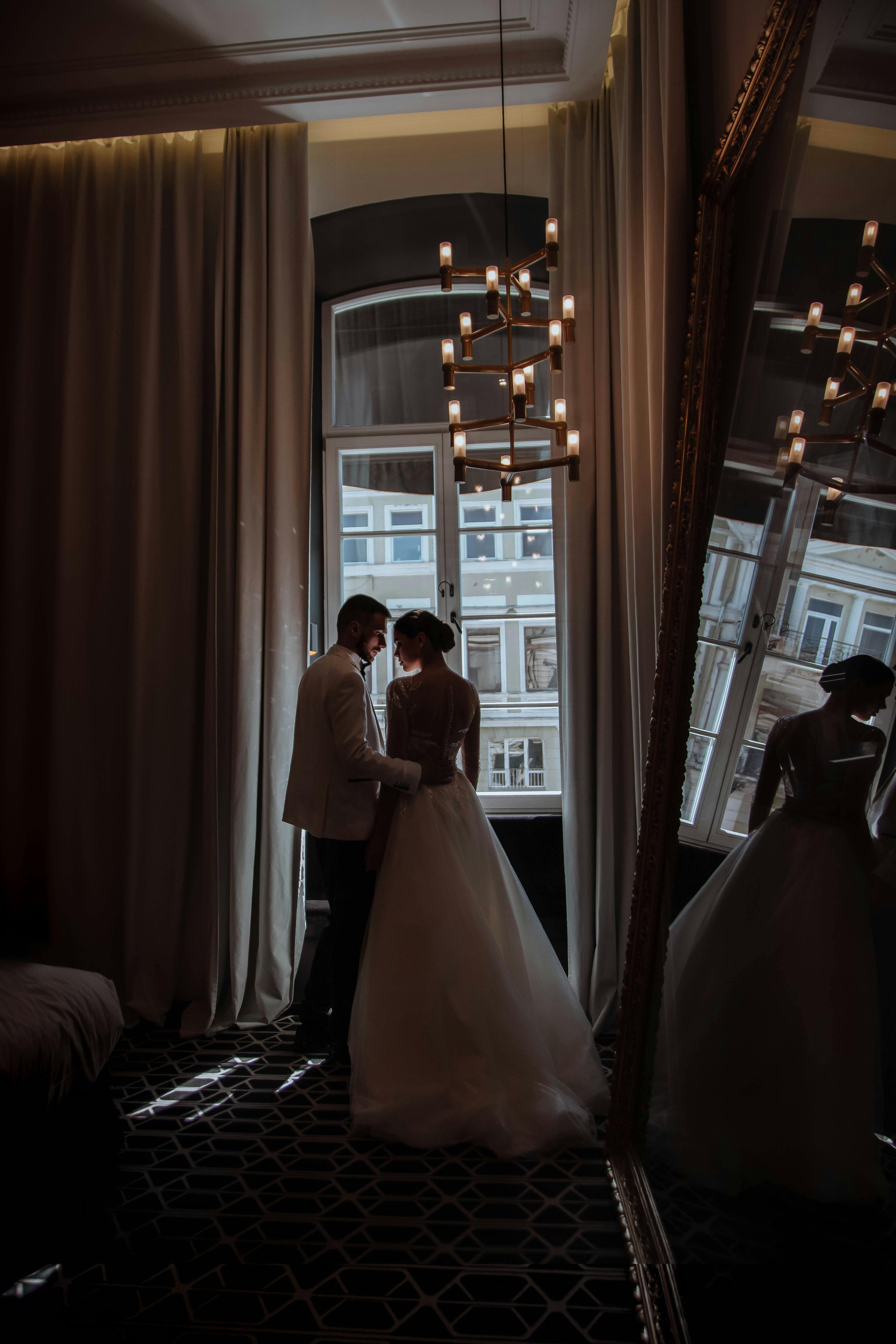 A bride and groom sharing a moment in a dimly lit room with large windows and ornate decor, their silhouettes reflected in a tall mirror.