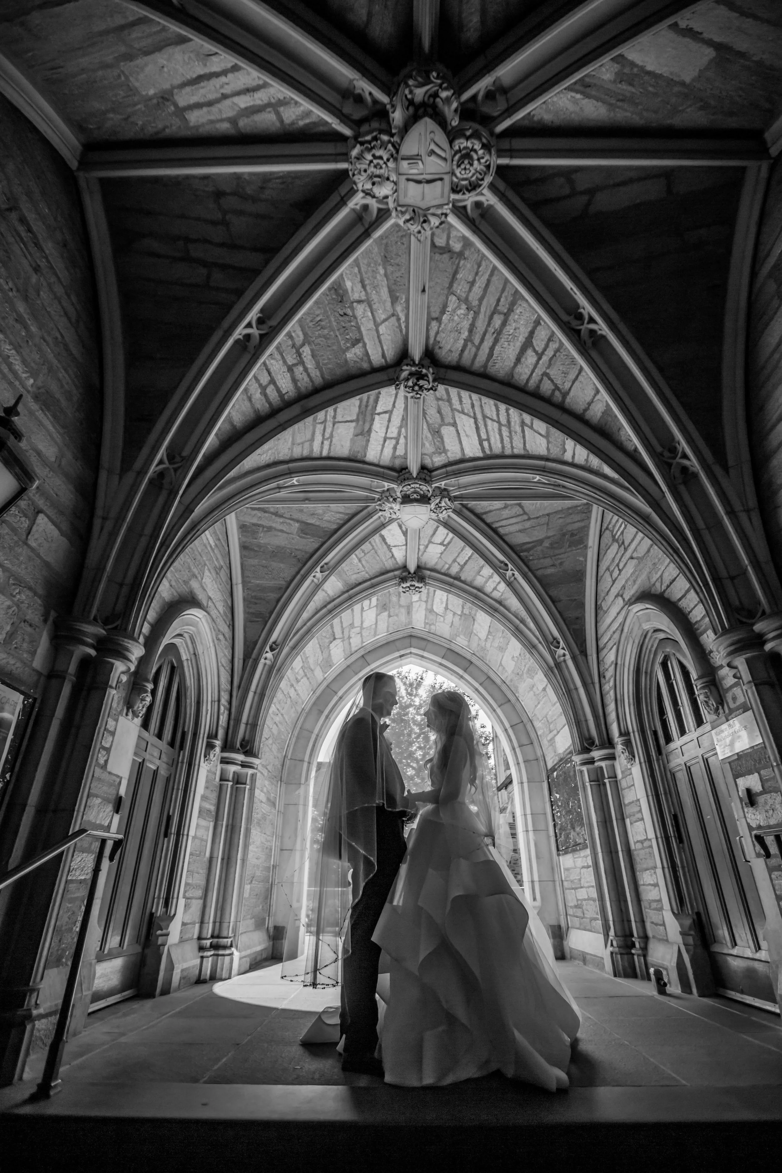 Bride and groom holding hands inside a Gothic-style stone archway, with natural light through a window behind them.